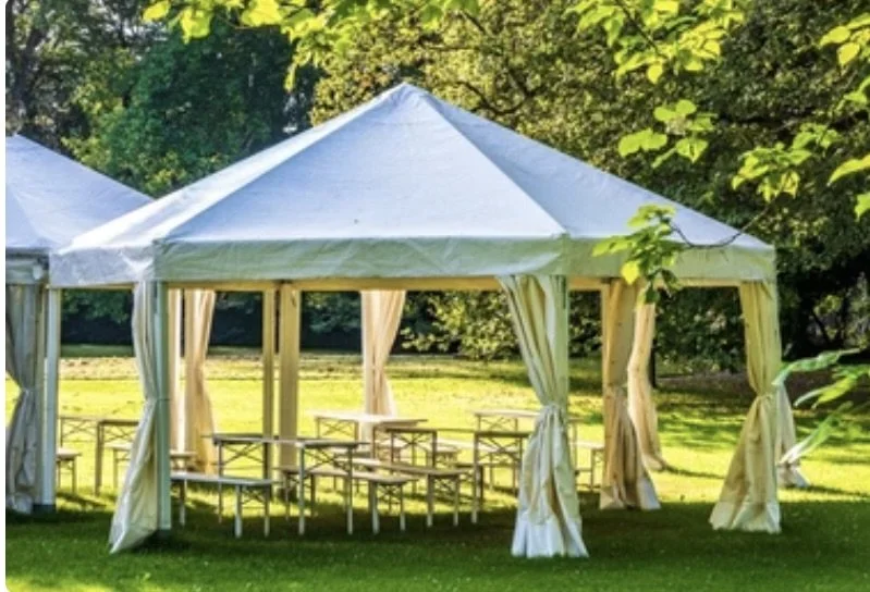 White outdoor event tent with open sides and draped curtains, set up on a grassy area with trees in the background.