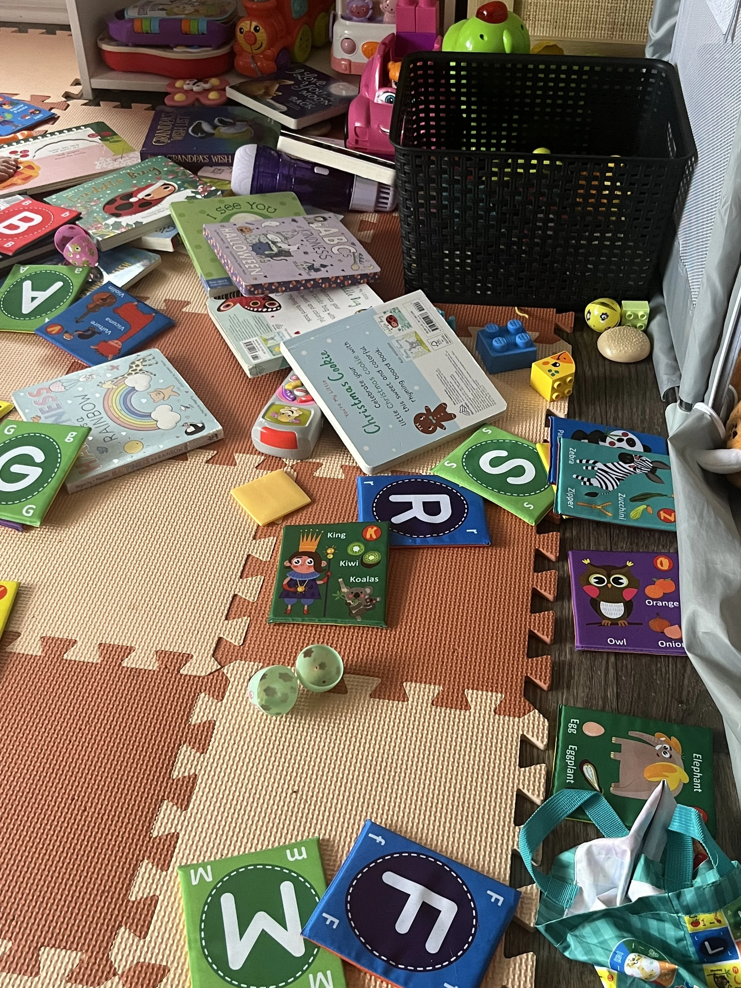 A messy play area with scattered children's books, toys, blocks, and educational cards on the floor and a black storage basket filled with more toys.
