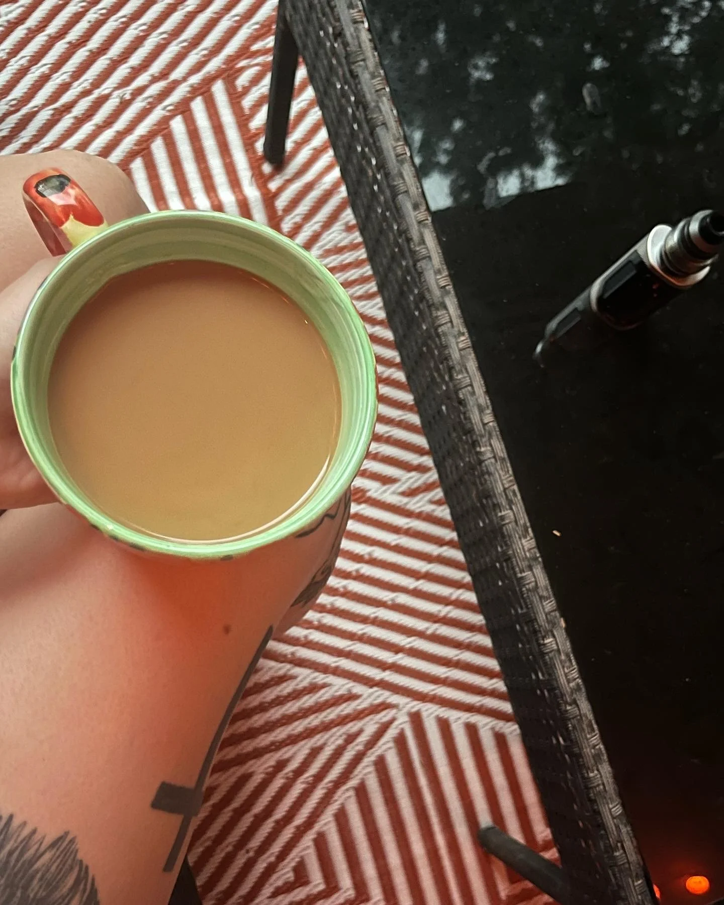 A person's hand holding a mug of coffee or tea with cream, resting on their knee. The person has a tattoo on their arm and resides on a striped orange and white cushion. In the background, there is a glass-top table with an essential oil roller and a small brown bottle.