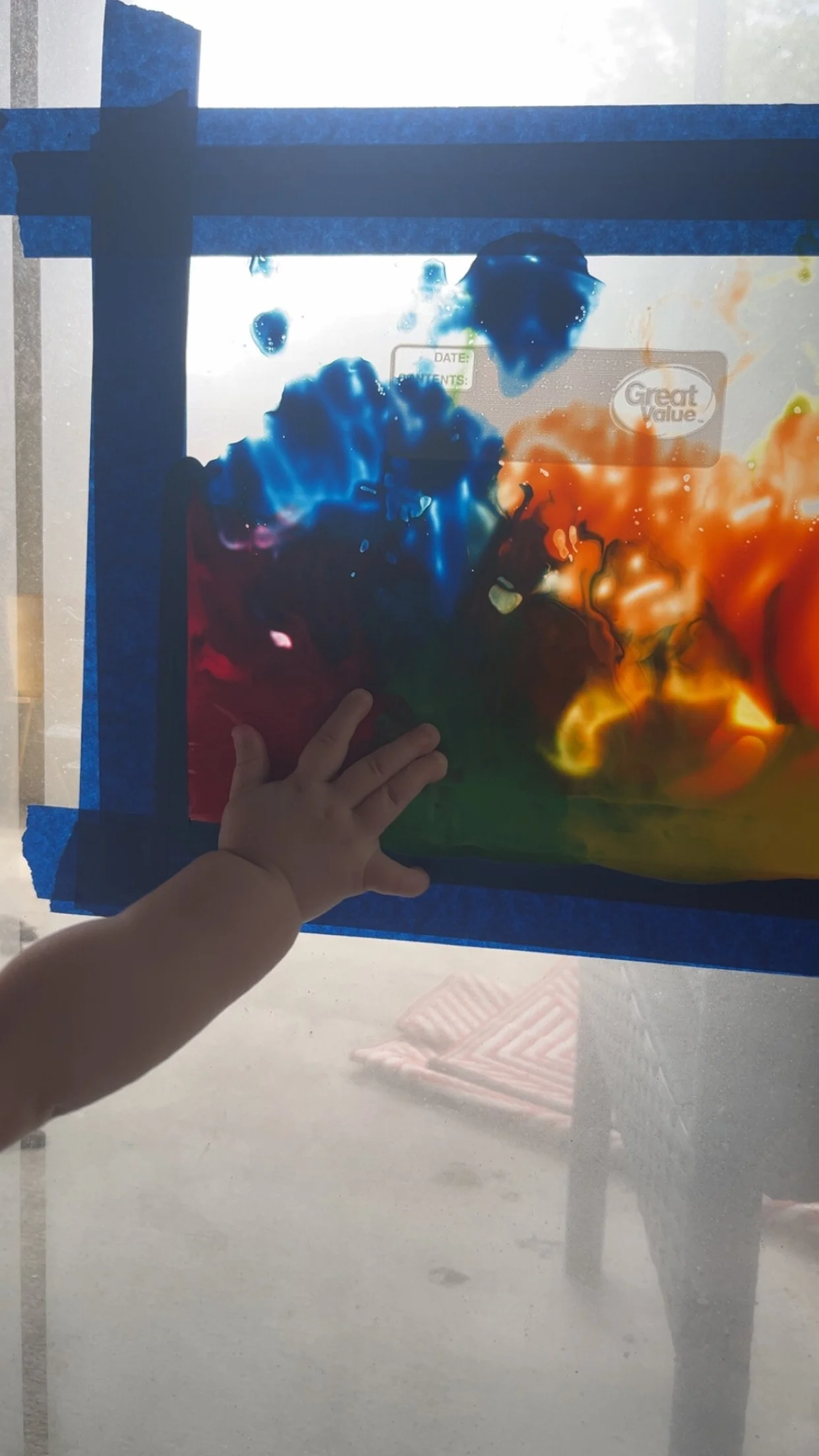 A child's hand touching a colorful abstract painting on a glass surface, with blue tape framing the artwork and a reflection of chairs and floor in the background.