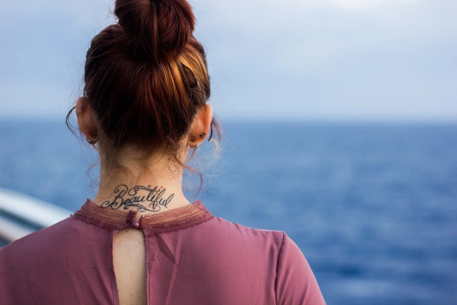 Back of a woman with red hair in a bun, wearing a pink top, standing outdoors by the ocean, with a tattoo that reads 'Beautiful' on her neck.