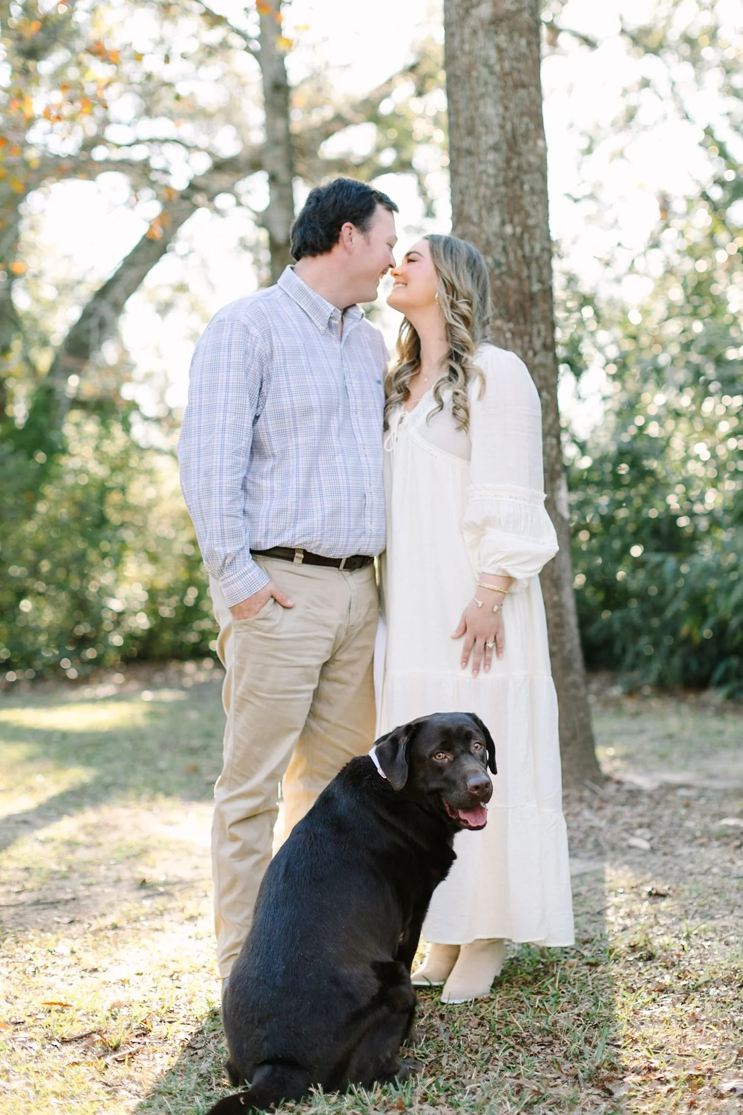 A man and woman are smiling and touching noses in a wooded outdoor setting with sunlight filtering through trees, and a black dog sitting in front of them.