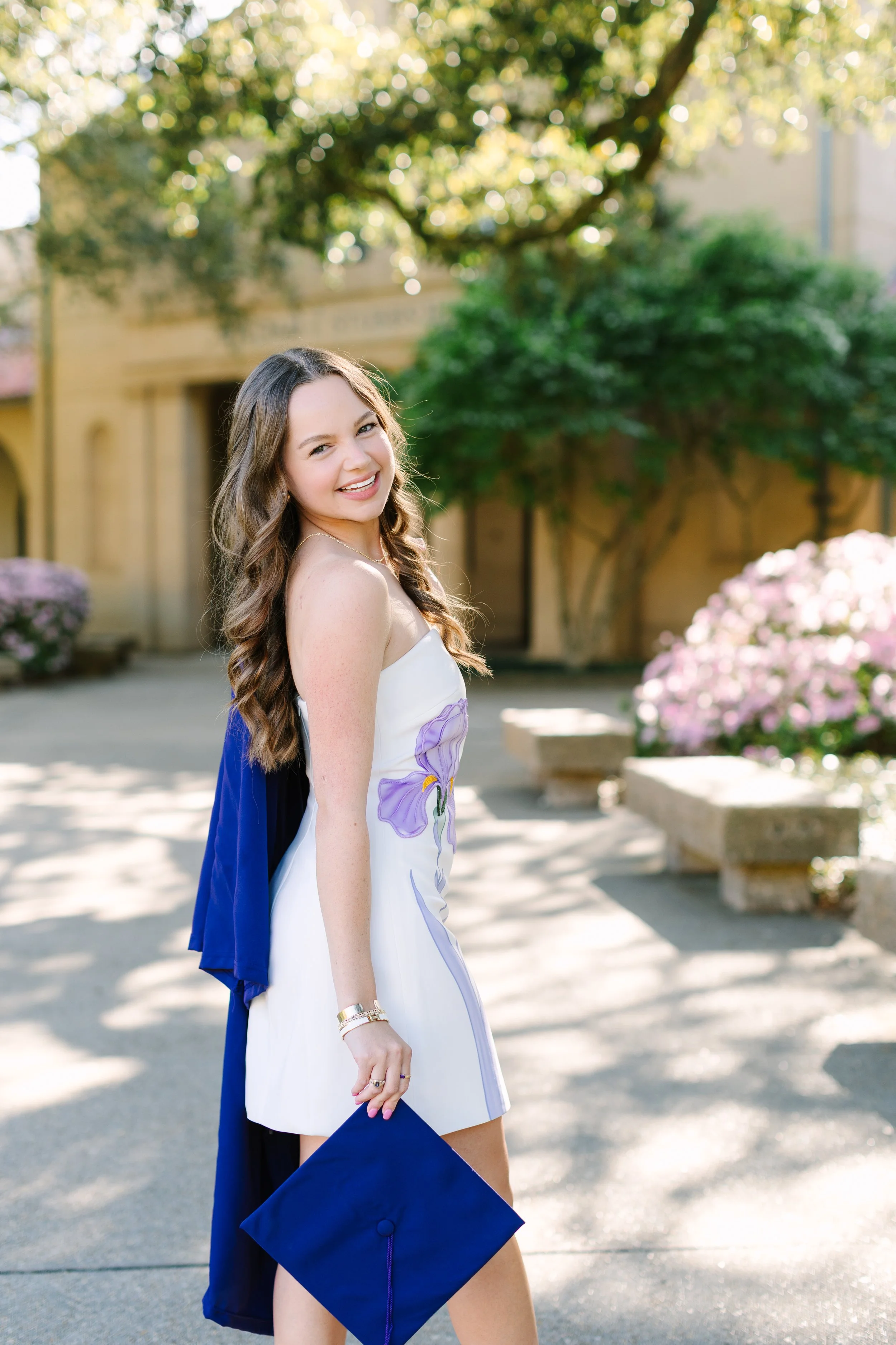 A young woman celebrating graduation outdoors, holding a blue diploma cover, smiling, with trees and flowers in the background.