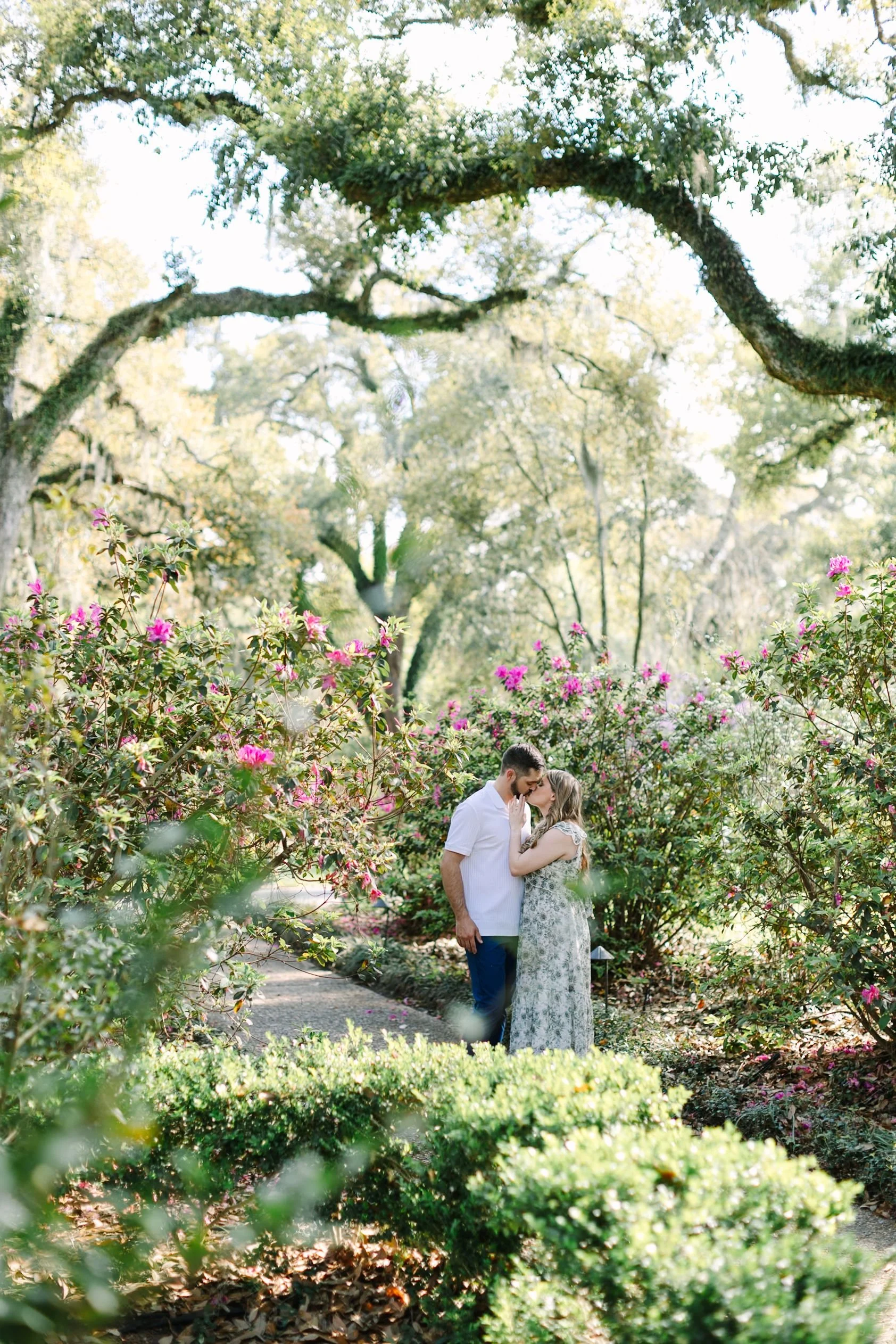 A couple sharing a kiss in a lush garden with pink flowers and large trees.
