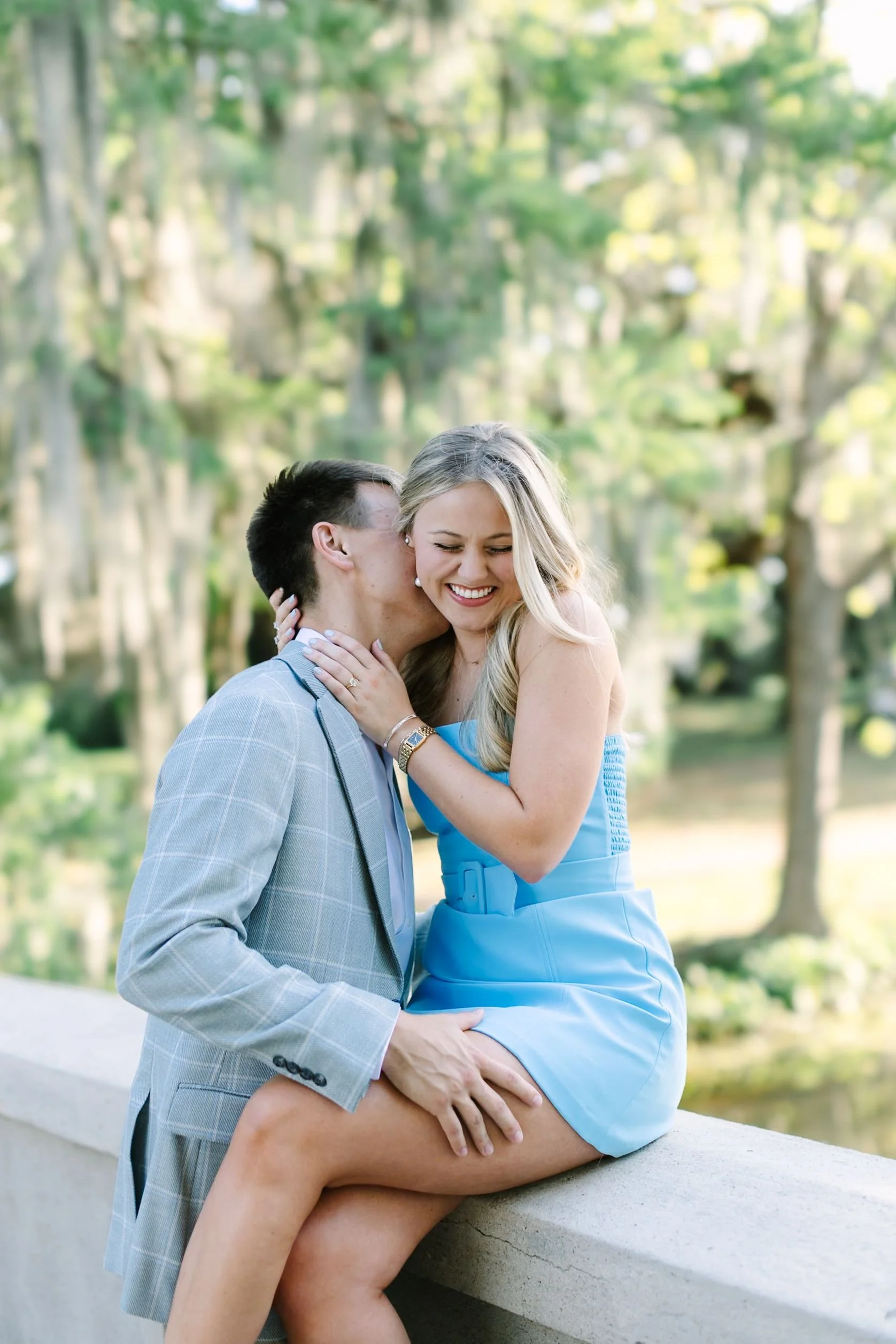 A couple sitting on a low wall outdoors, sharing an intimate kiss and smiling, with the woman wearing a sleeveless blue dress and the man in a light gray suit, surrounded by a lush green park in the background.