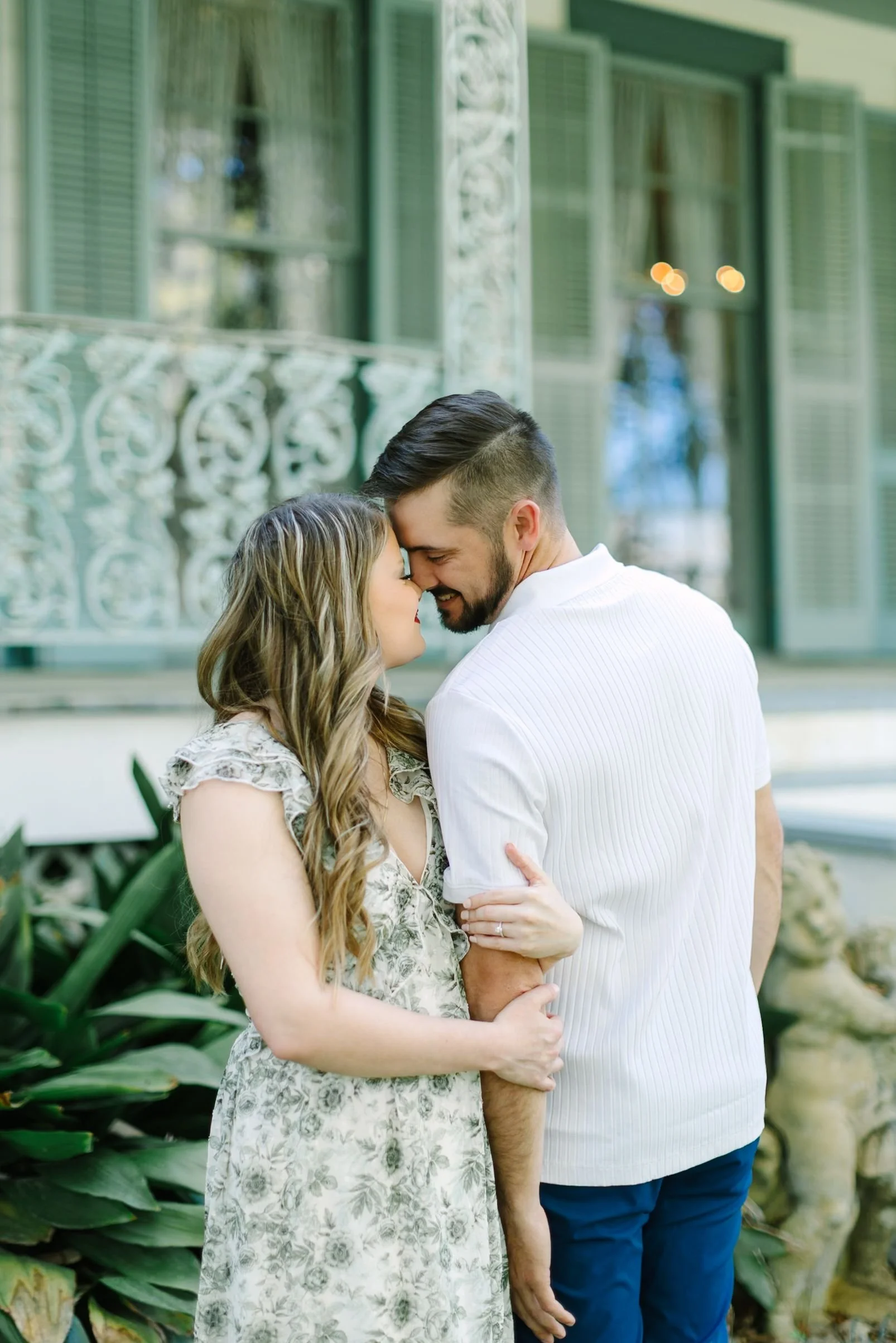 A couple shares a tender moment with foreheads touching, standing close outdoors surrounded by greenery and a vintage house with a wrought iron balcony.