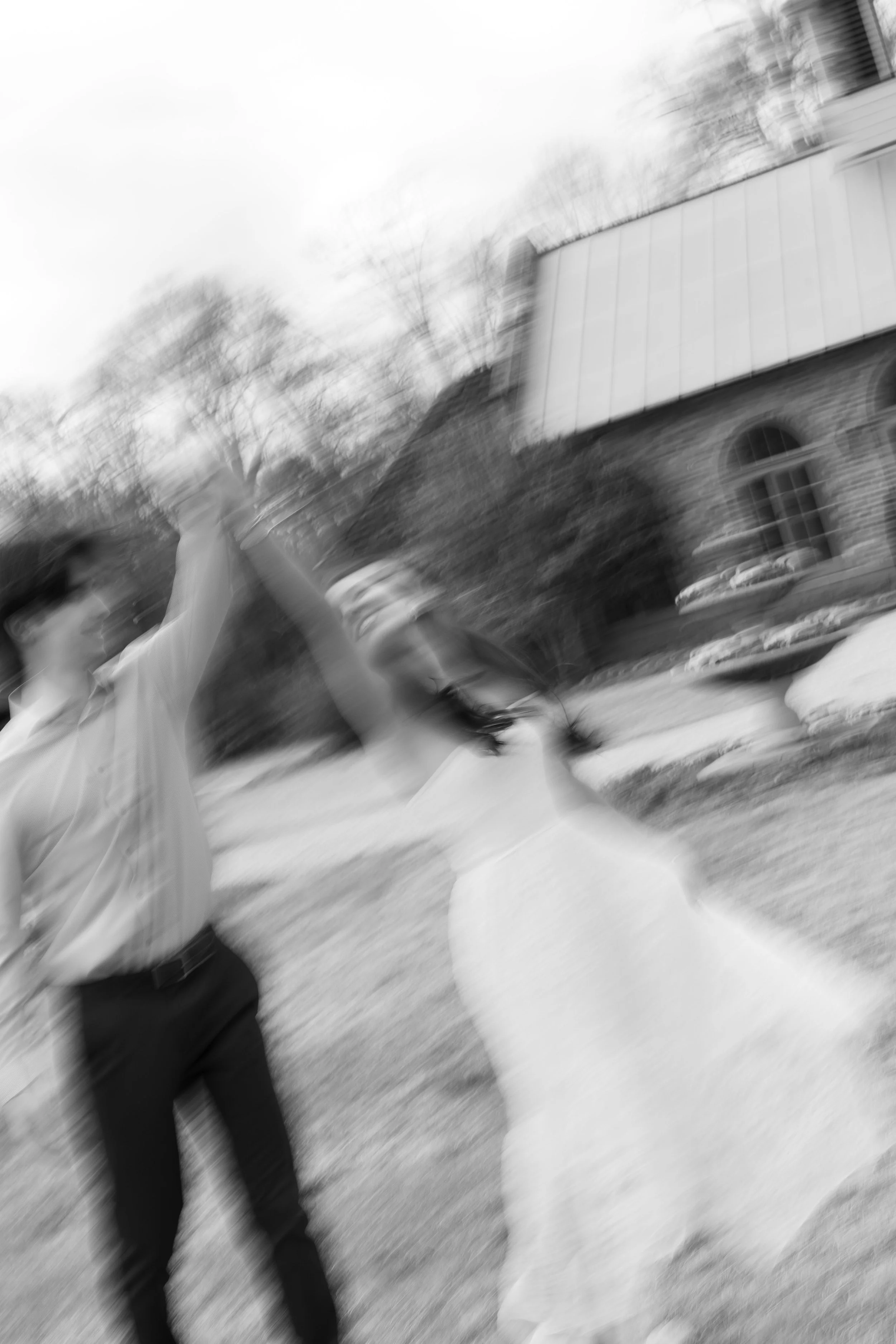 A blurry black and white photo of a couple dancing outdoors near a house, with trees in the background.