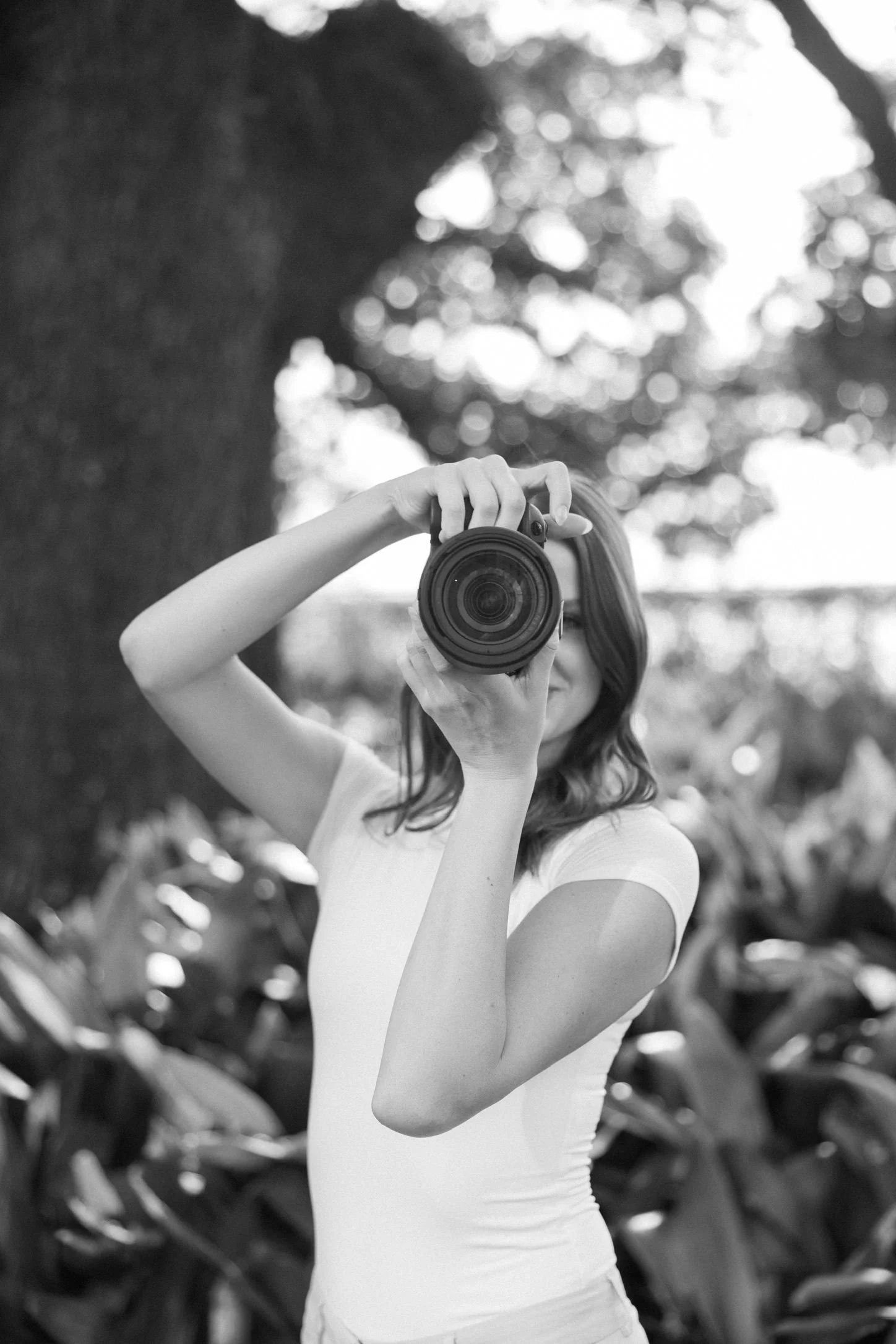 A woman with shoulder-length hair wearing a white t-shirt, taking a photo with a camera outdoors. She is standing in front of a background with a large tree and blurred plants, and is facing the camera while holding it up to her face.