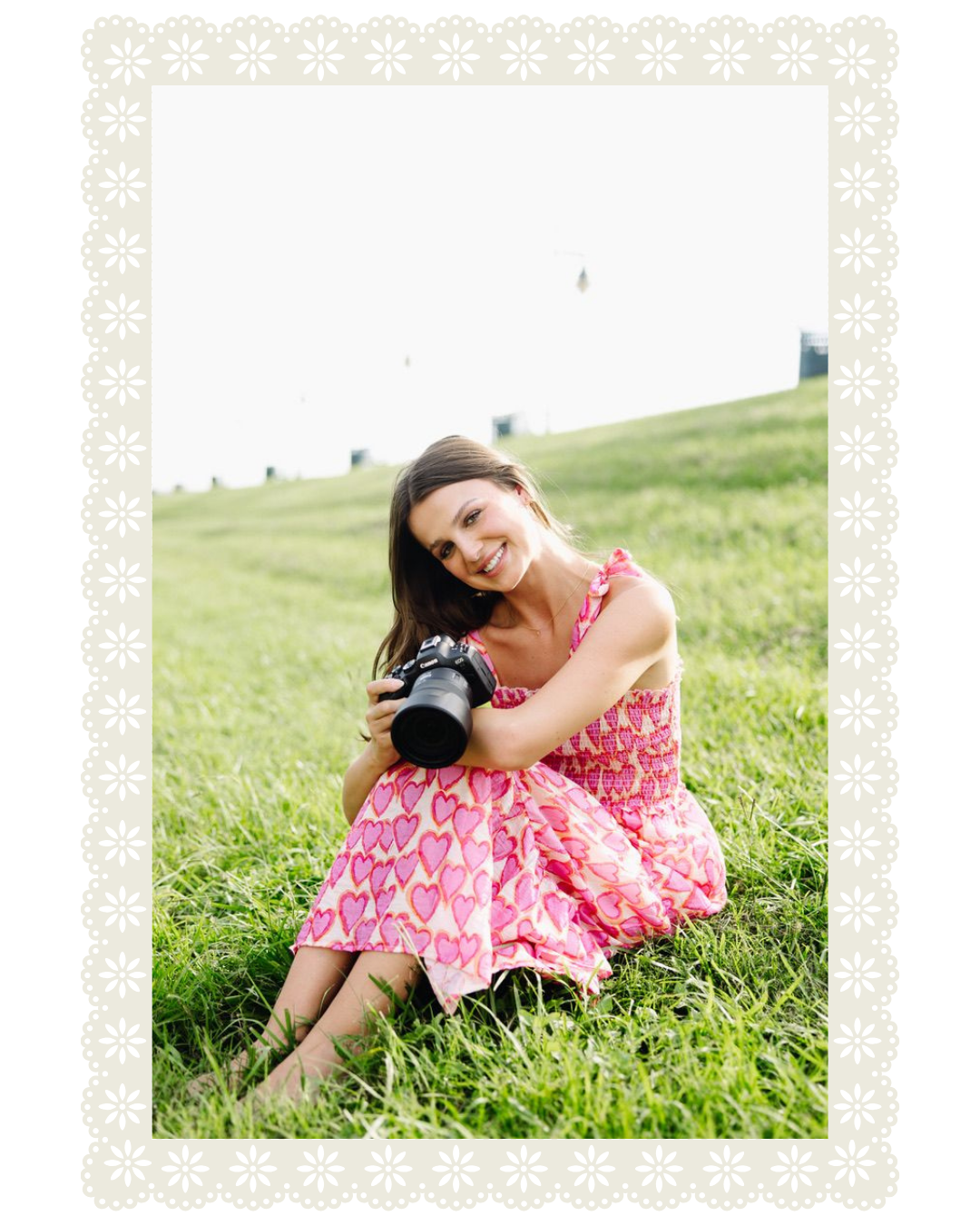 A smiling woman sitting on grass, holding a camera, wearing a pink dress with heart patterns, outdoors on a bright day.