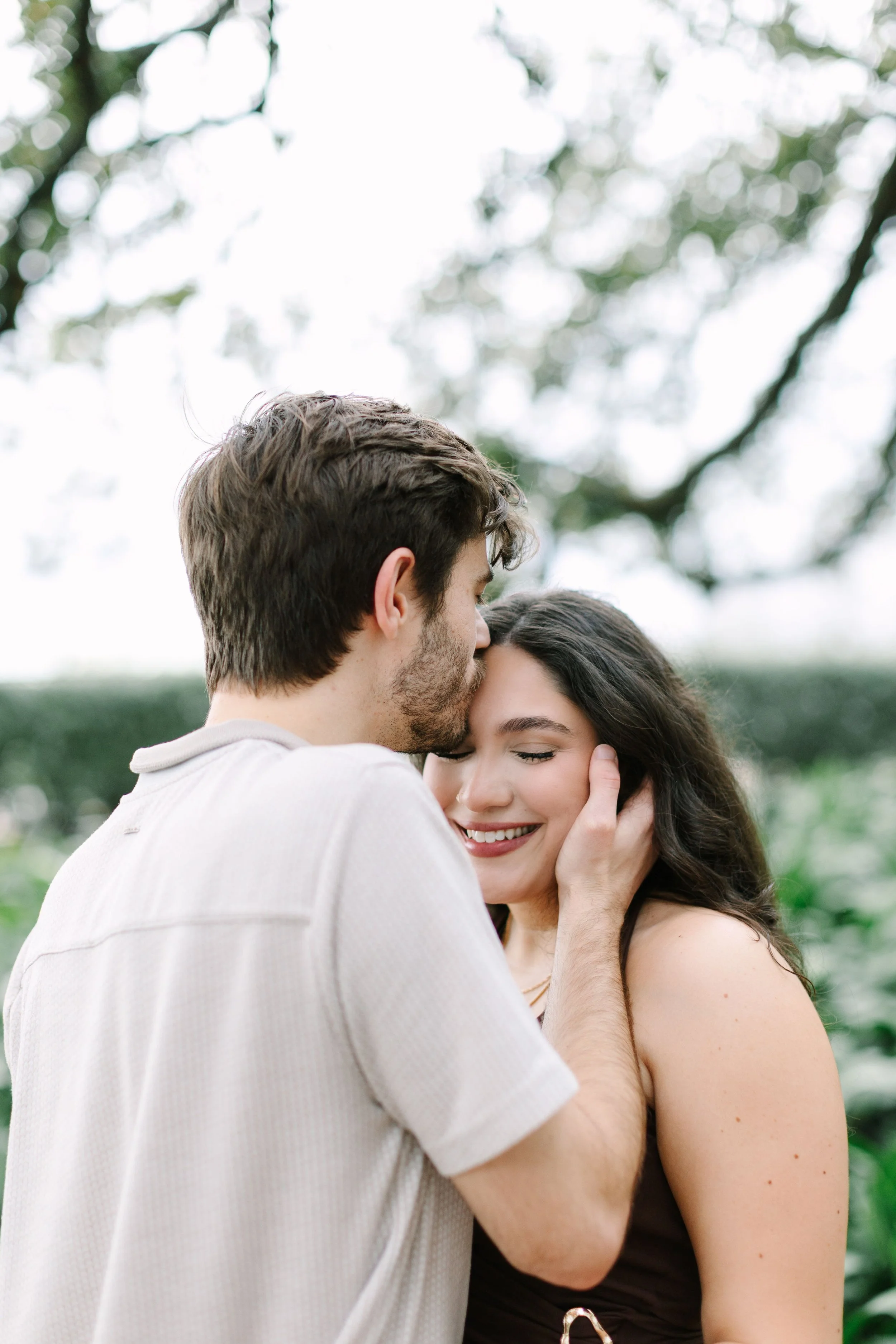 A young couple sharing an intimate moment outdoors, with the man gently holding the woman's face and whispering to her, both smiling.