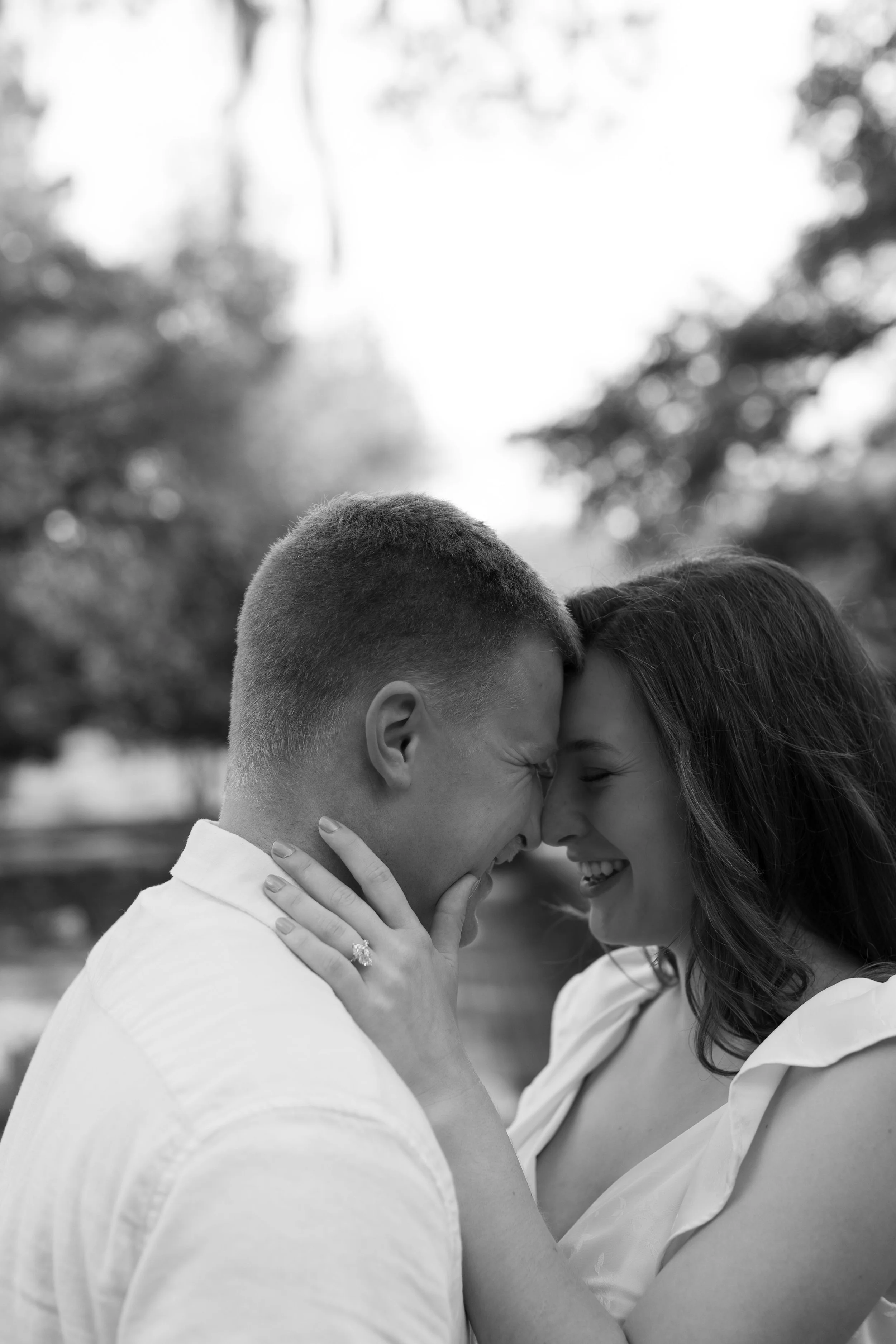 A black and white photograph of a smiling couple with their foreheads touching outdoors, with trees and blurred background.