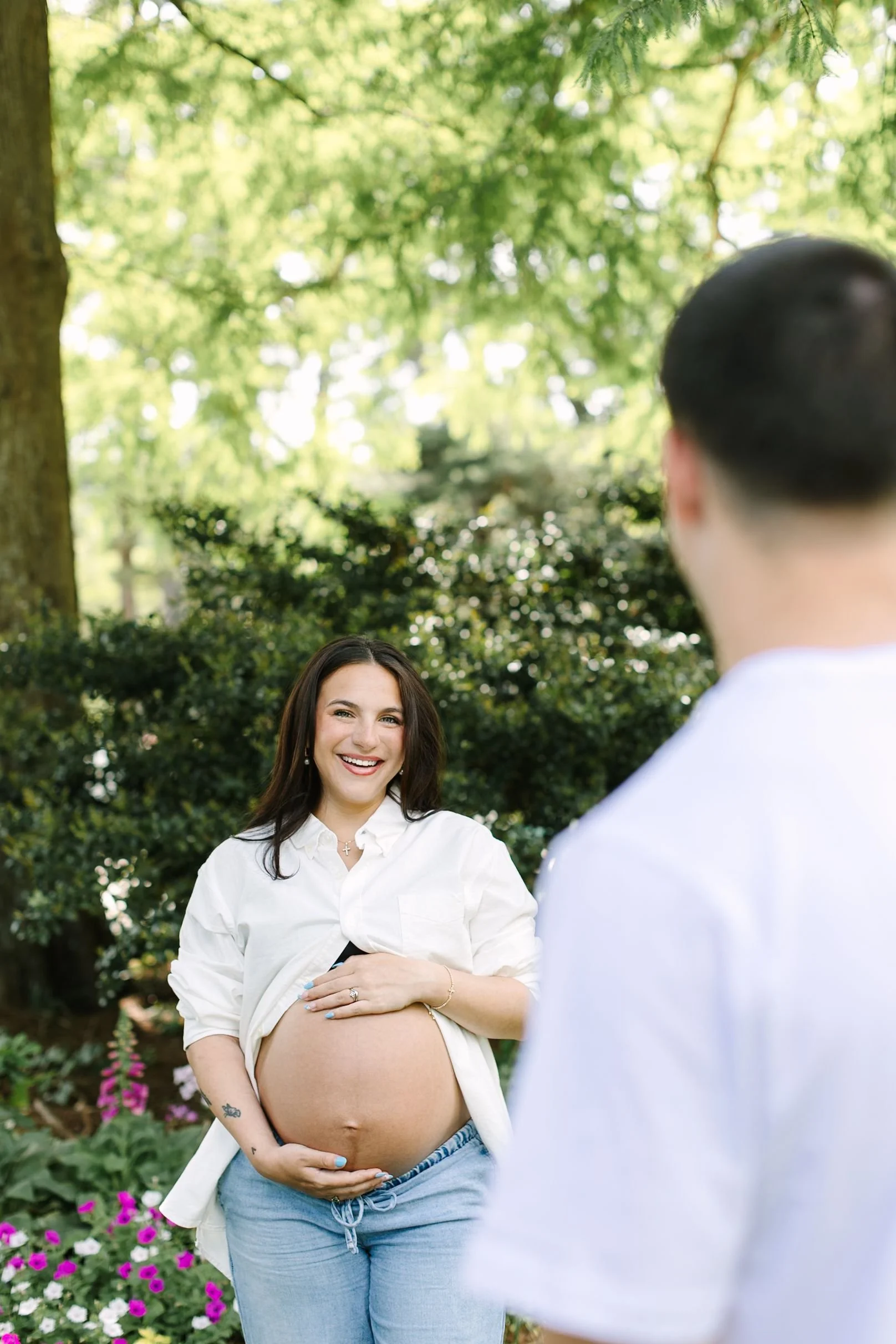 A pregnant woman smiling and holding her belly outdoors, with a man facing her, in a lush garden with green trees and colorful flowers.