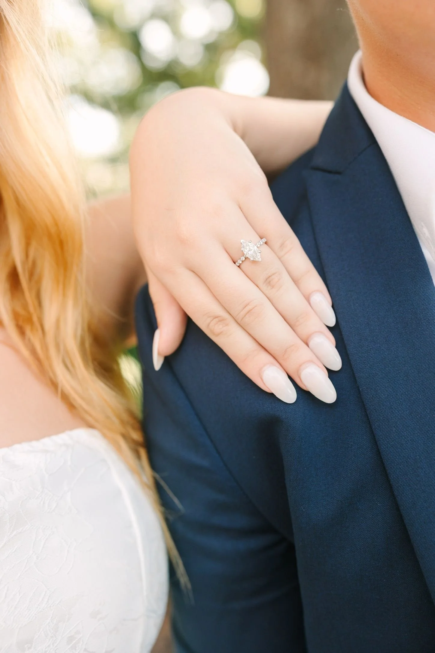 A woman with a diamond engagement ring resting her hand on a man's shoulder, who is wearing a navy blue suit and white shirt.