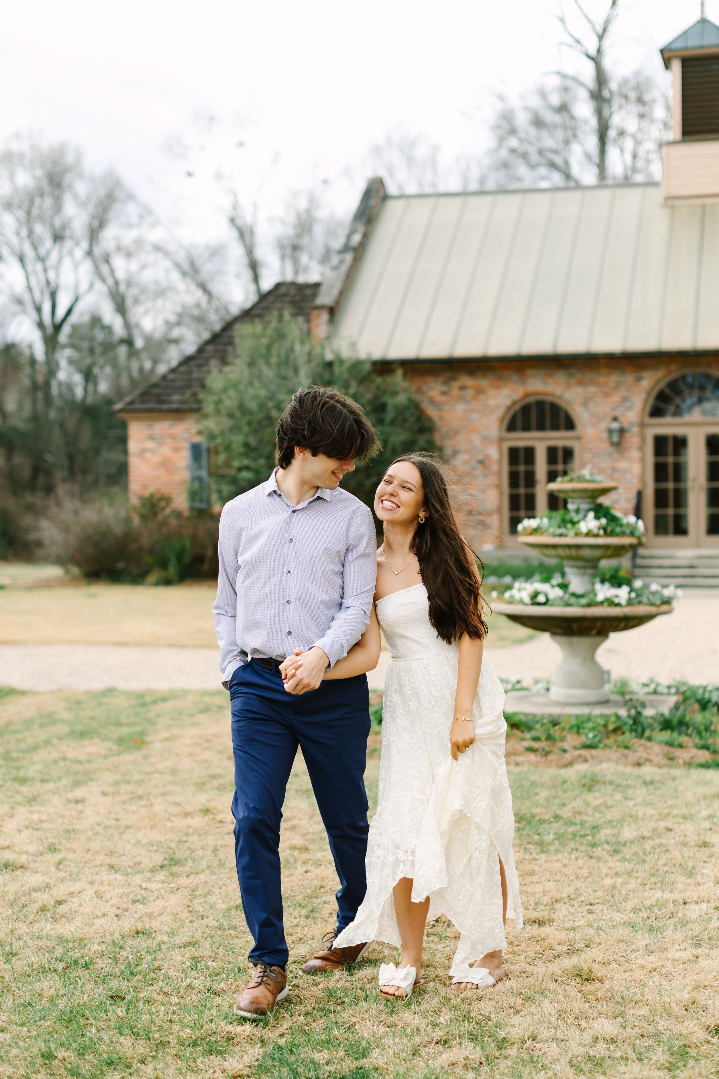 A happy couple is walking together outside on a lawn in front of a brick house with a fountain and trees in the background.