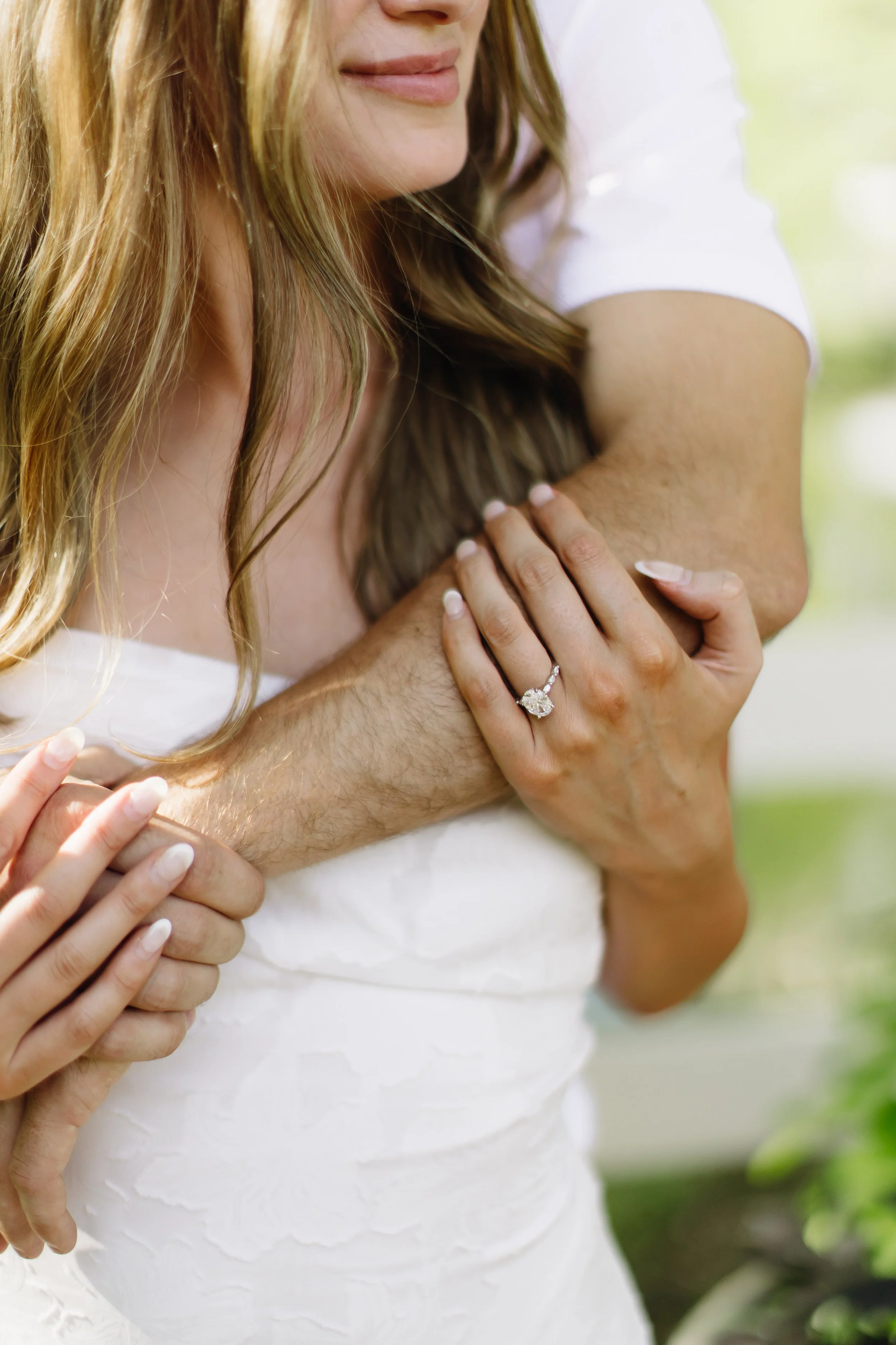 Close-up of a woman with long hair wearing a white dress, showing an engagement ring on her finger, with a man behind her. They are outdoors with greenery in the background.