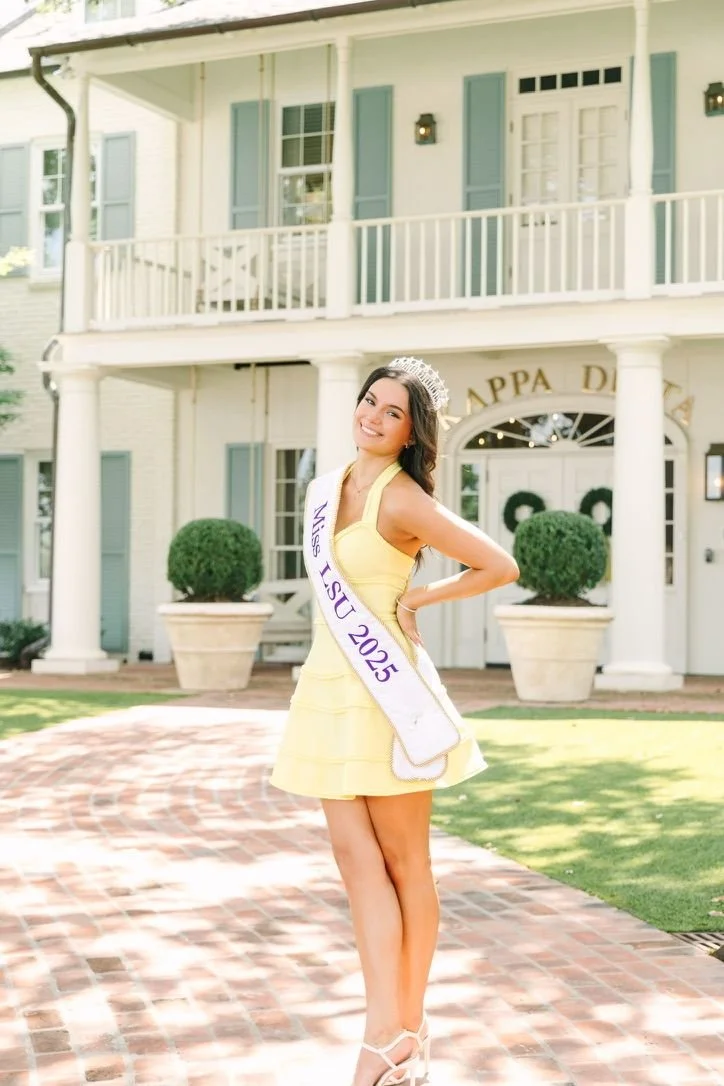Miss LSU 2025 standing outside a house with a white porch, wearing a yellow dress, a sash reading "Miss LSU 2025," a tiara, and high heels, smiling at the camera.