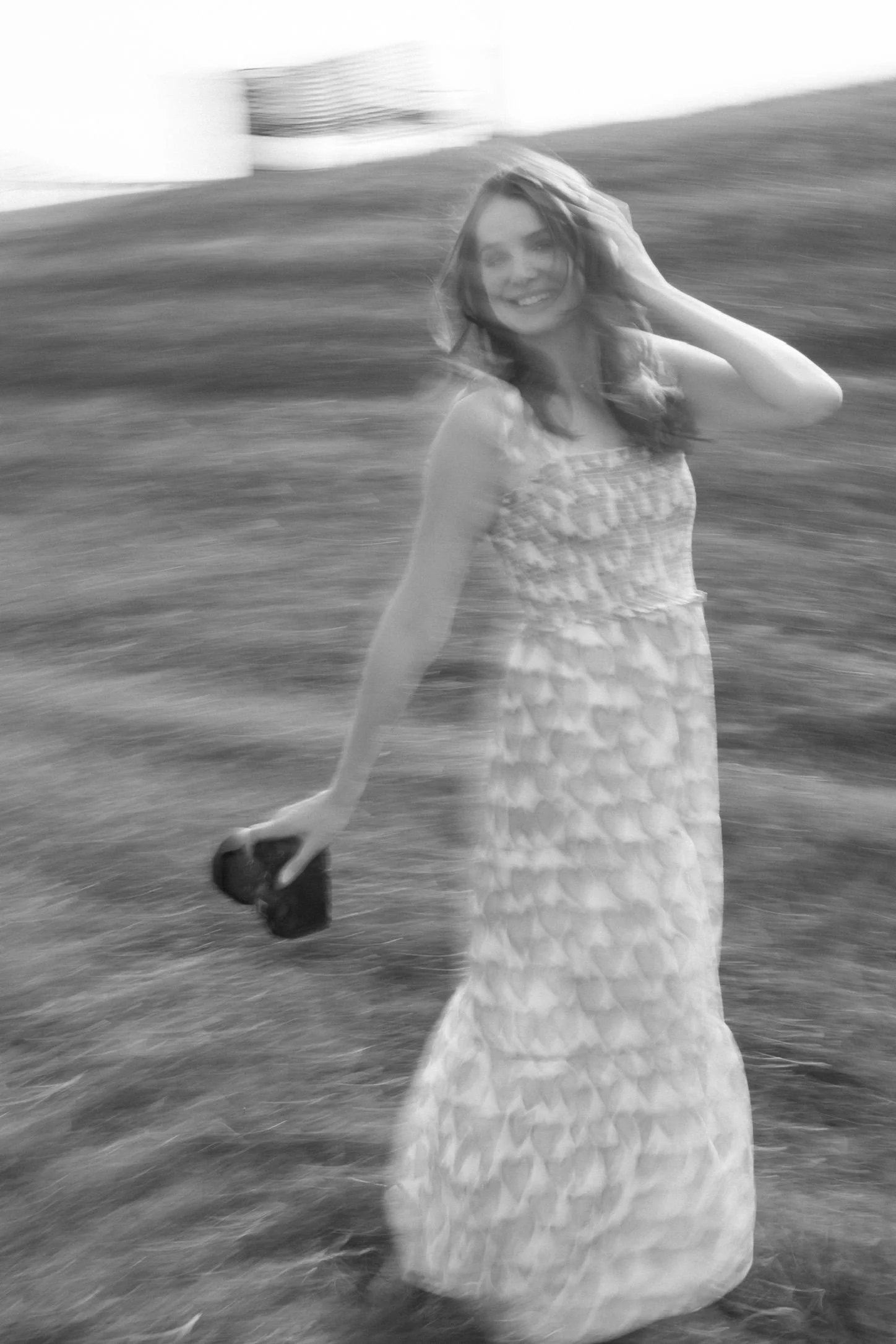 Black and white photo of a smiling young woman in a patterned dress holding a camera, standing outdoors on grass, with motion blur background and motion effect