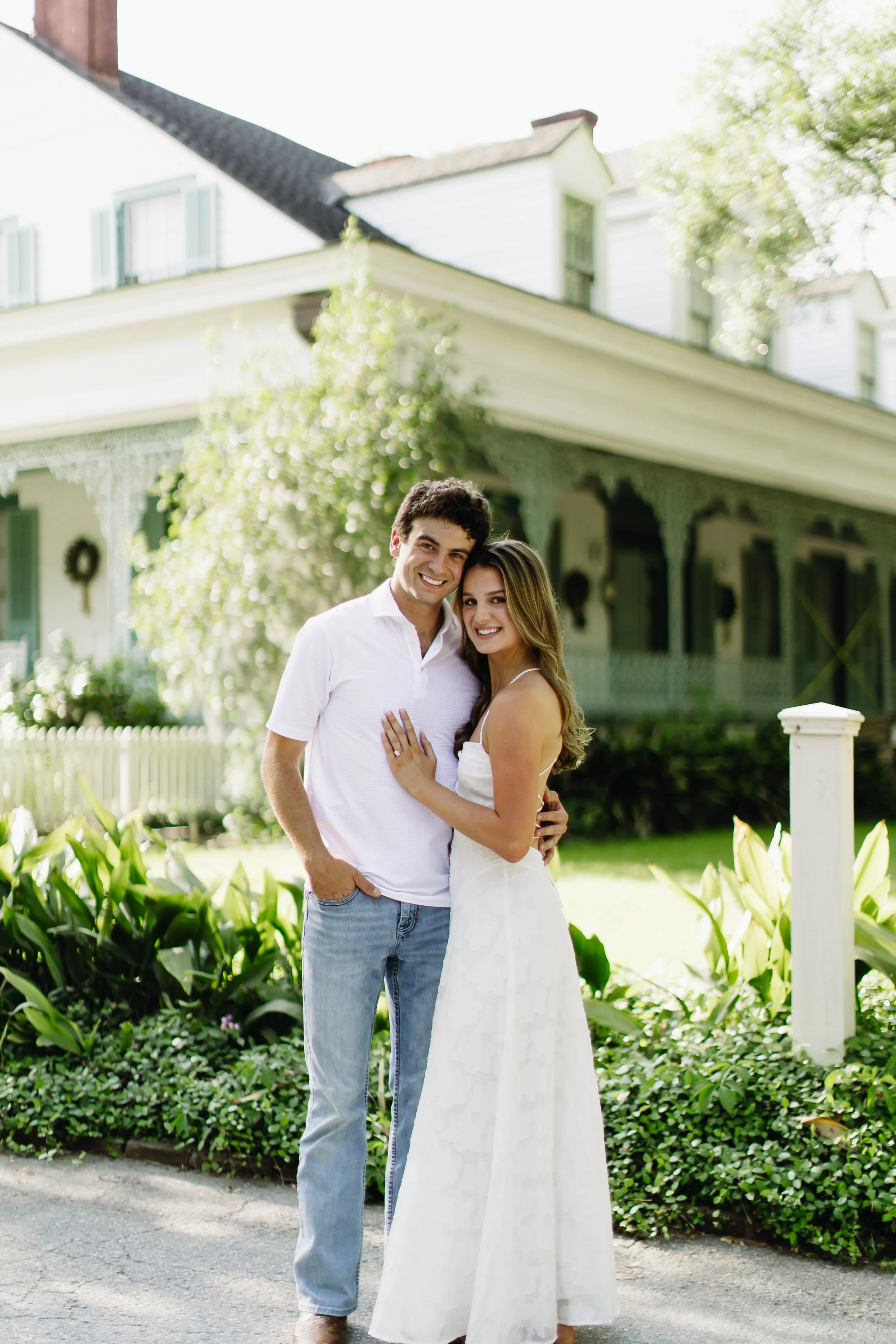 A smiling young couple stands close together in front of a white house with green shutters, surrounded by lush greenery and large white lilies.
