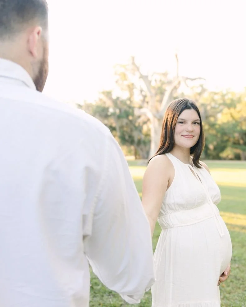 A pregnant woman in a white dress smiling at the camera while holding her belly, with a man in a white shirt holding her hand, outdoors on a sunny day with trees in the background.
