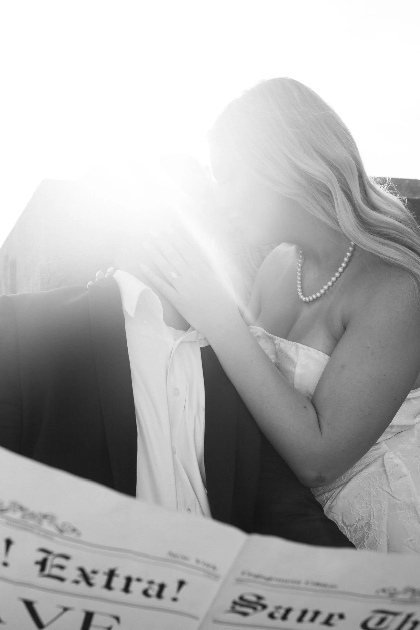 A woman in a wedding dress and pearl necklace kissing a man dressed in a tuxedo, with sunlight shining behind them.
