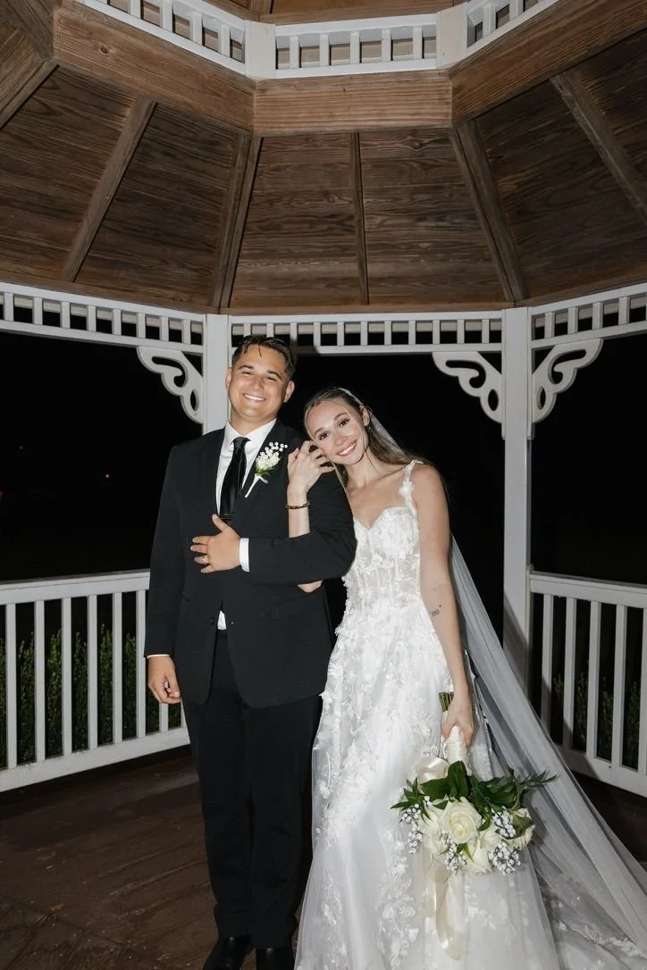 A newlywed couple stands in a gazebo at night, smiling and posing for a photo. The groom is wearing a black tuxedo with a white shirt and black tie, and the bride is in a white lace wedding gown holding a bouquet of white roses with greenery.