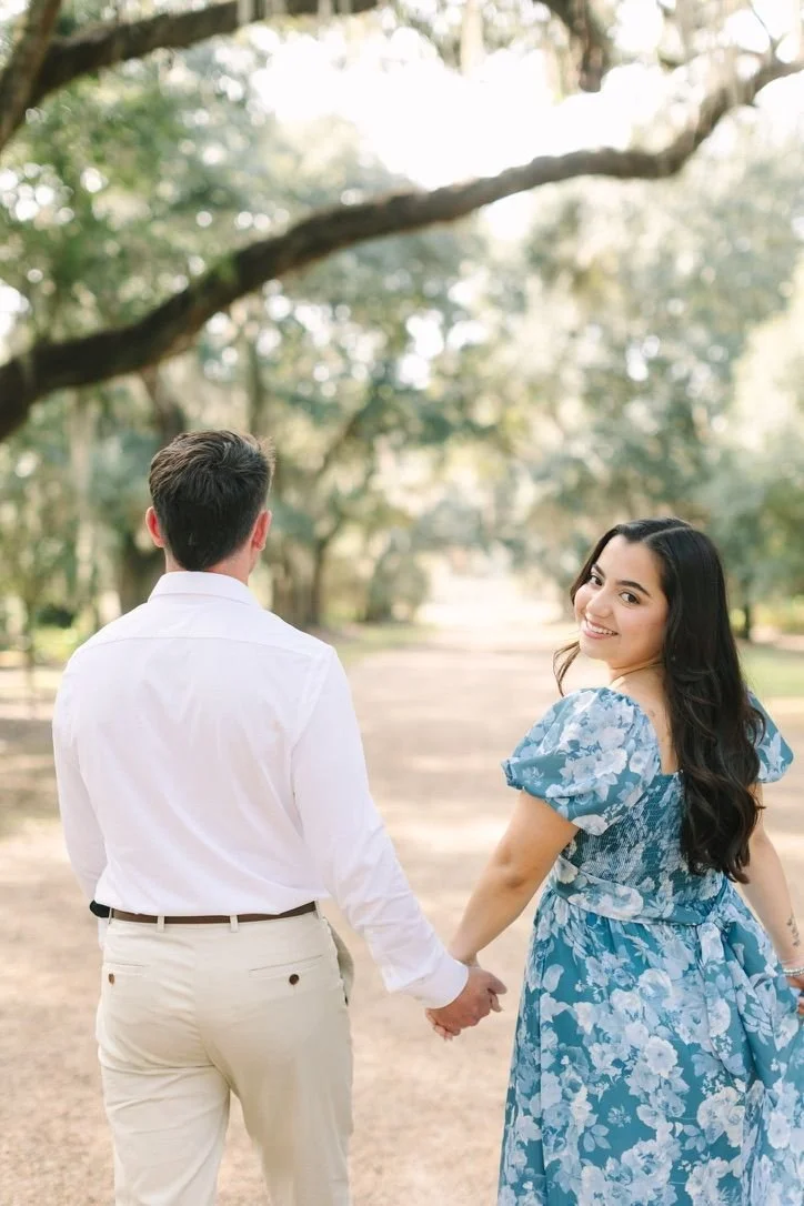 A young couple holding hands in a park, with the woman smiling at the camera and the man facing away, surrounded by green trees and sunlight.