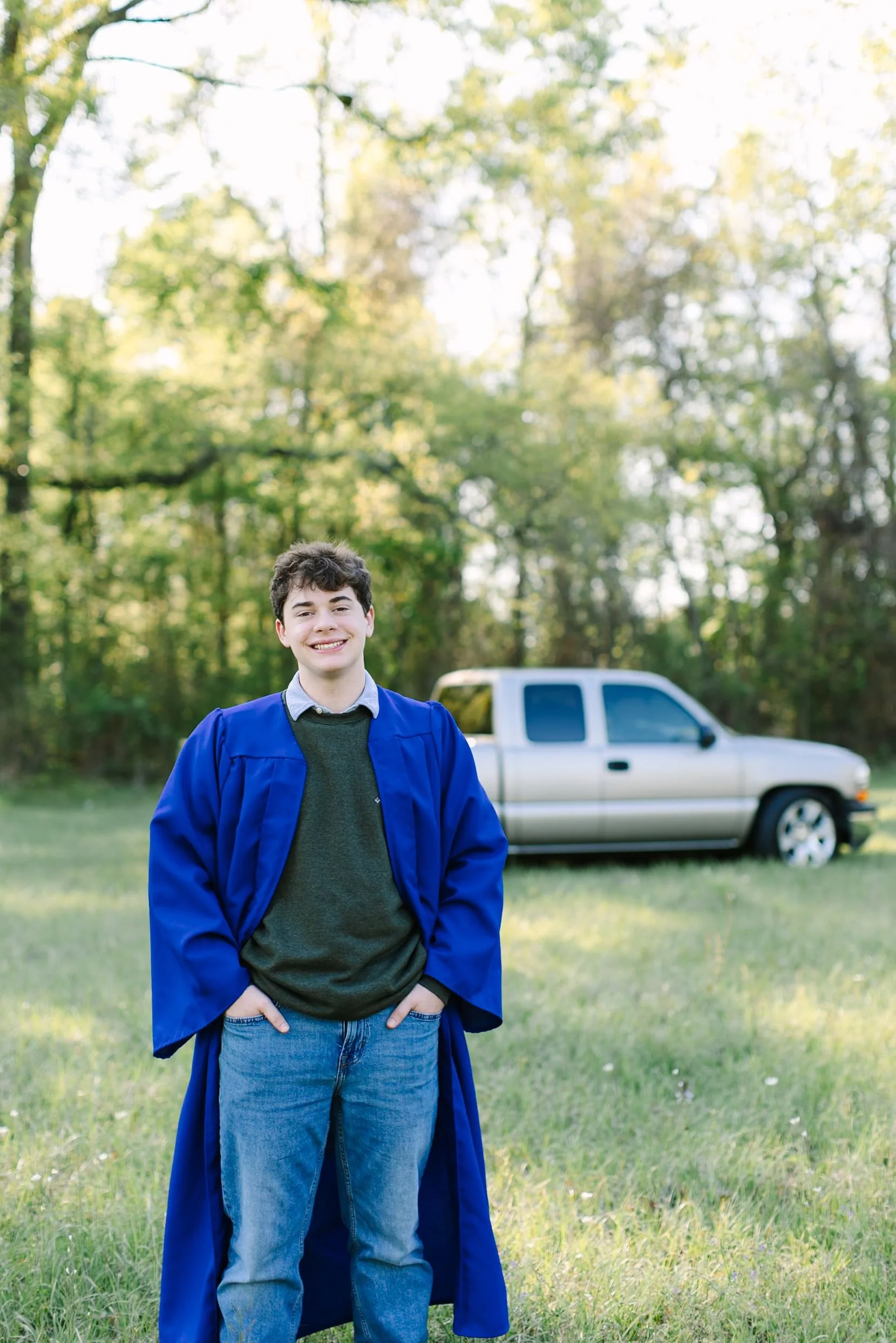 A teenage boy in a blue graduation gown standing in a grassy field with a silver pickup truck behind him and trees in the background, smiling at the camera.