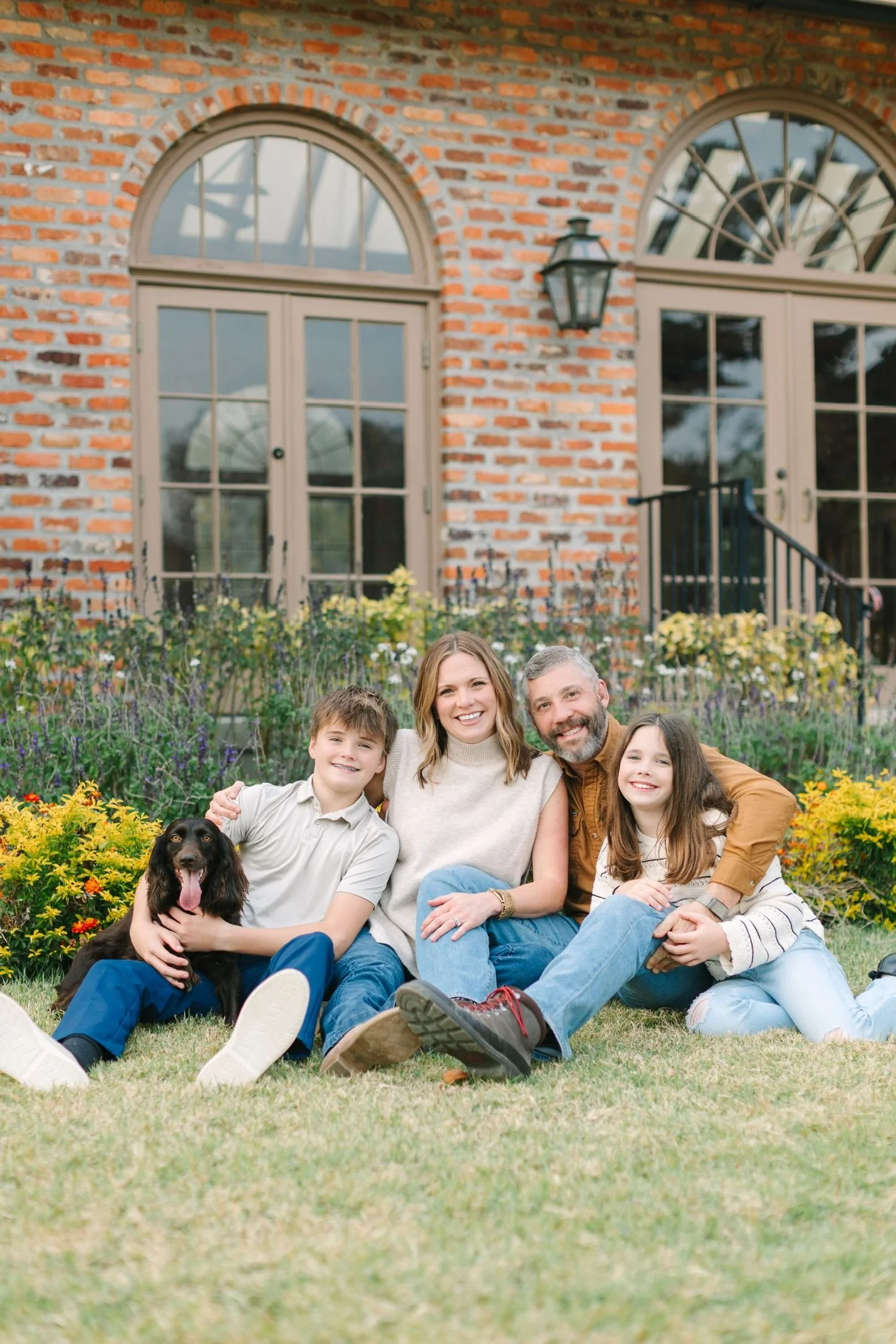 A family of four with a dog sitting on grass in front of a brick house with large arched windows and a garden.