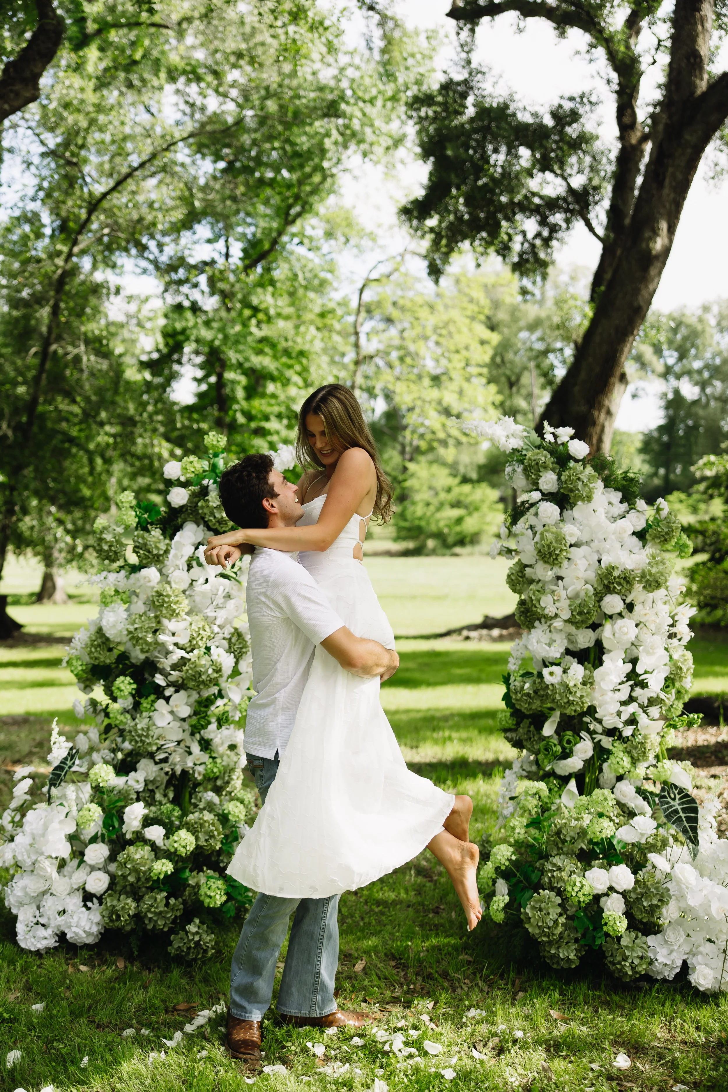 A man lifting a smiling woman in a white dress, surrounded by floral arrangements, in a lush green outdoor setting.