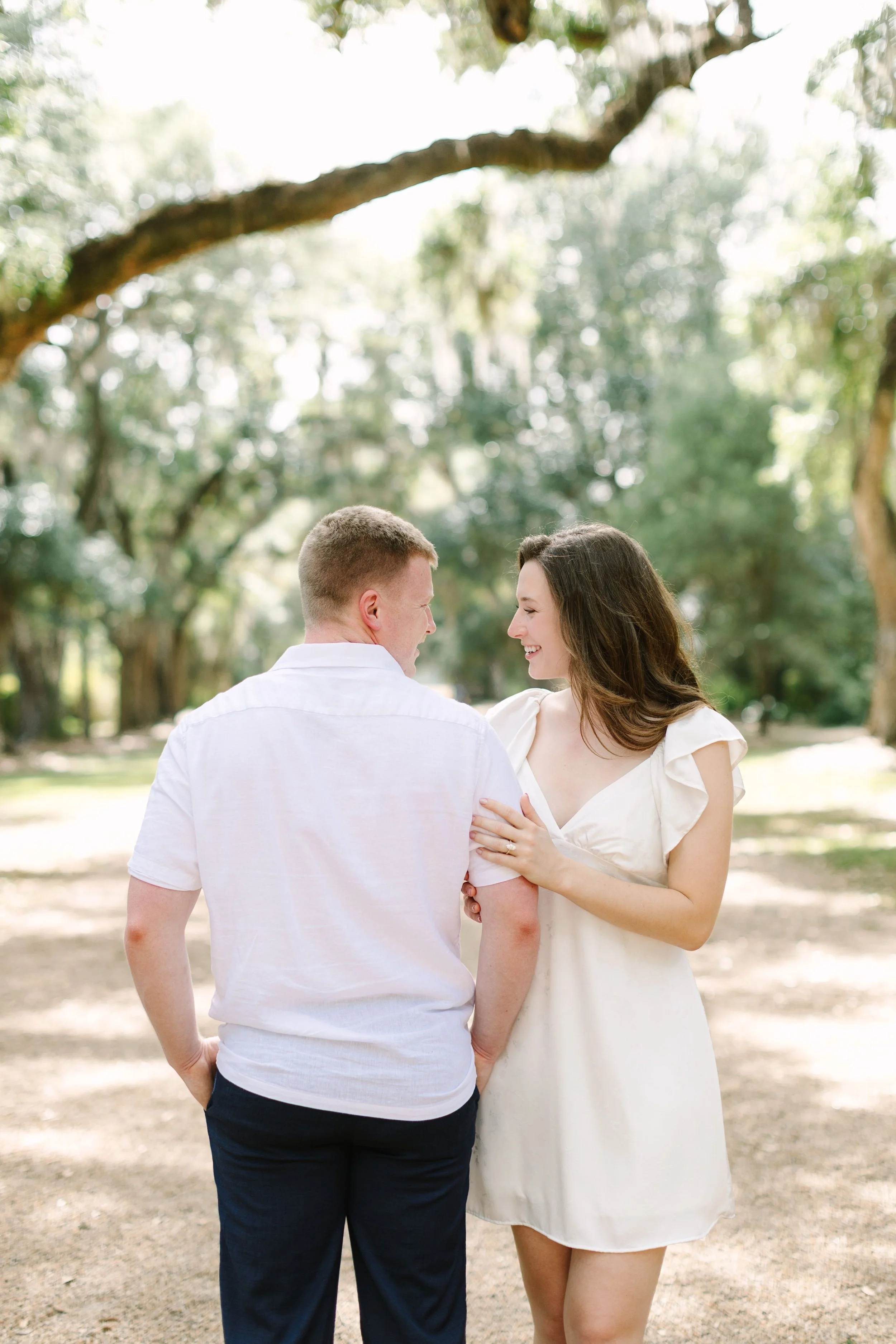 A man and woman standing outdoors in a wooded park, smiling at each other. The woman is wearing a white dress, and the man is in a white shirt and dark pants. They are close, with the woman's hand on the man's arm.