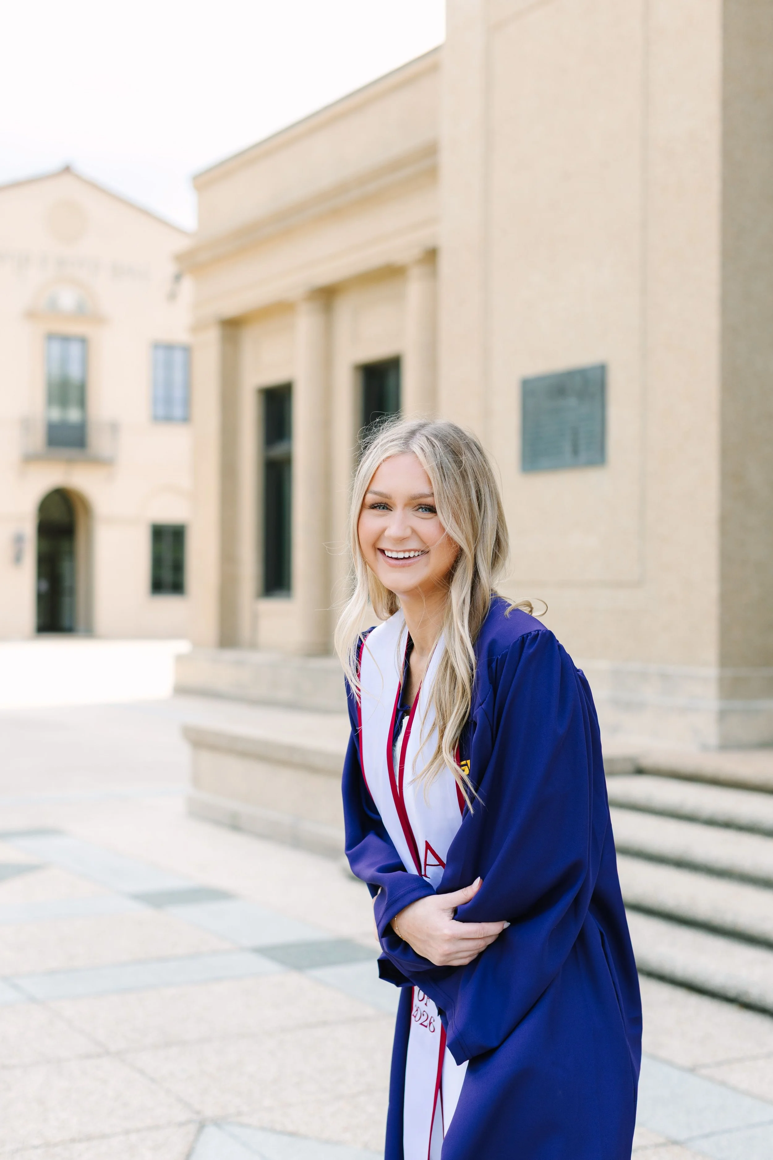 A young woman wearing a blue graduation gown and white sash, smiling outside a historic building with steps.