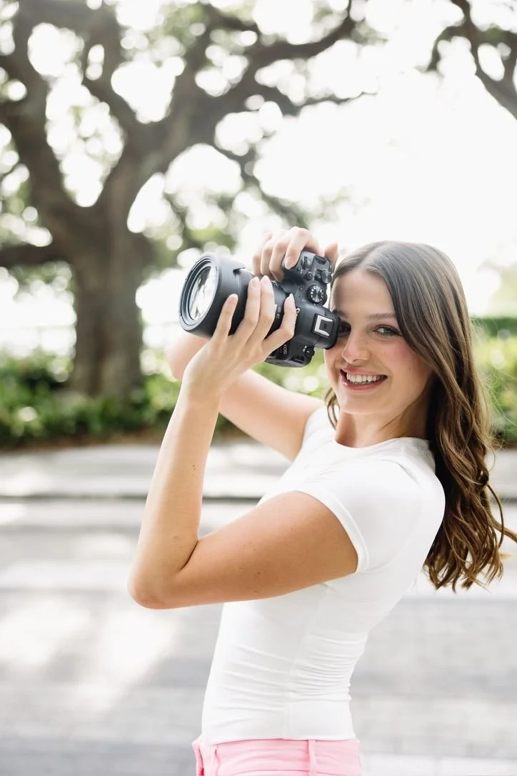 A woman with long brown hair smiling while holding a camera, outdoor setting with trees and sunlight in the background.
