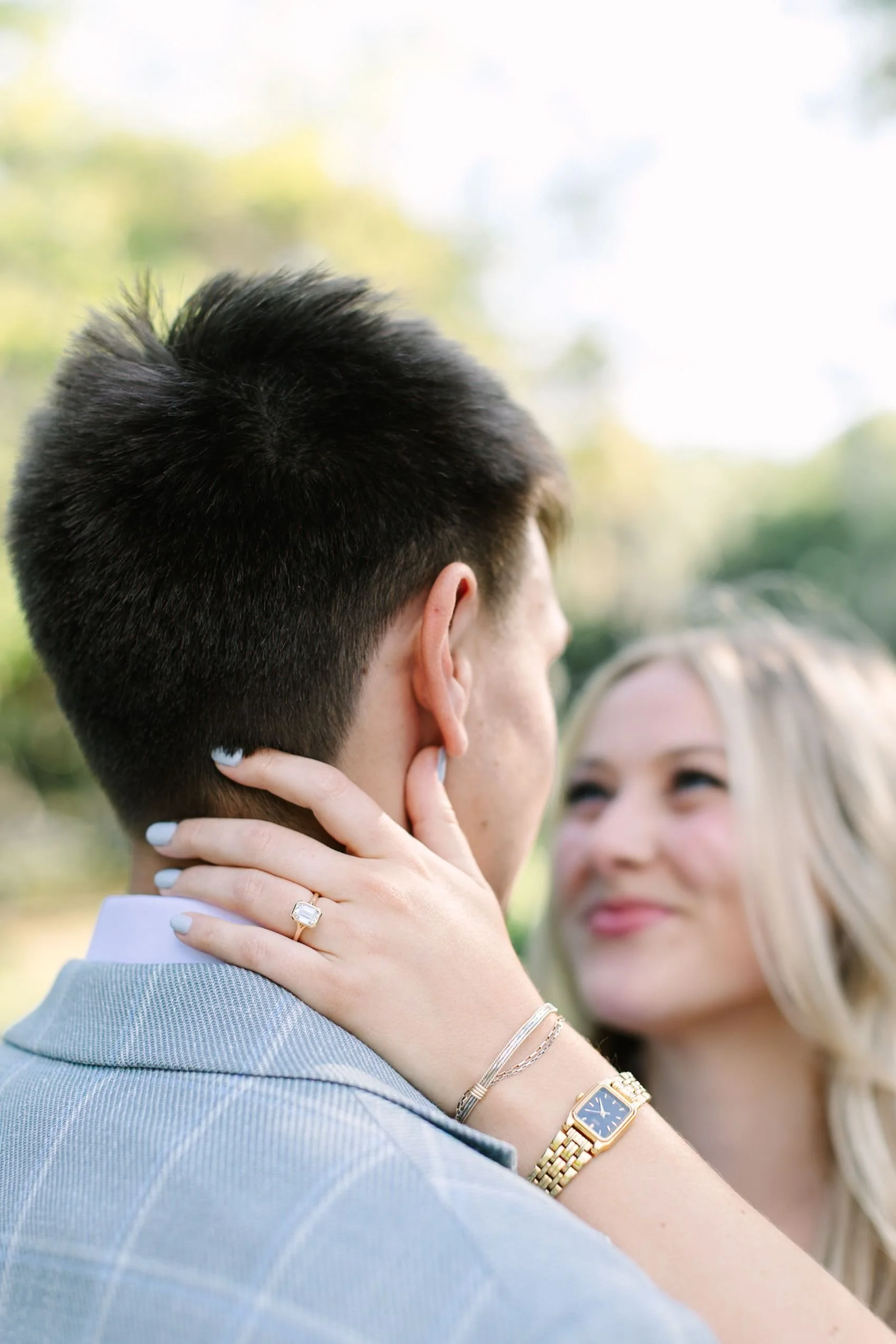 A close-up of a couple outdoors, woman touching man's face as they look at each other, with the woman wearing a gold watch, rings, and bracelets, and the man dressed in a light blue shirt with a checkered pattern.