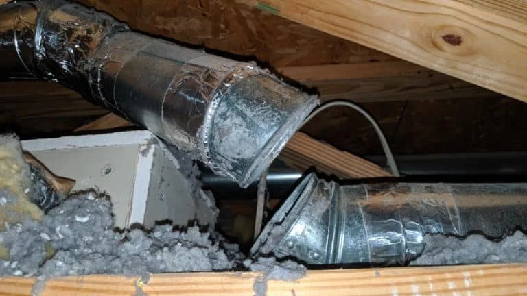 Close-up view of a damaged HVAC duct in an attic with insulation and wooden beams.
