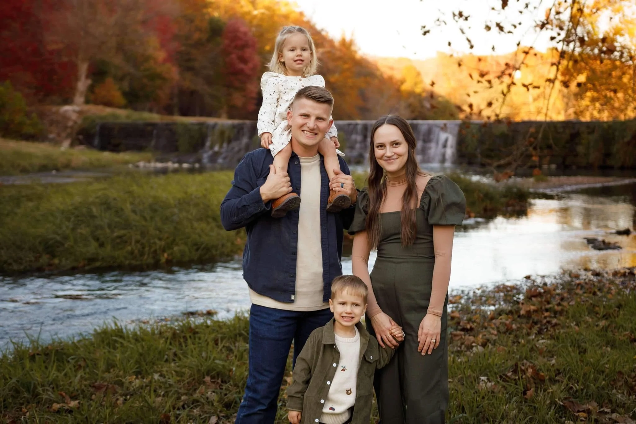 A family of five stands outdoors near a river with a waterfall in the background, surrounded by autumn trees with colorful fall foliage.