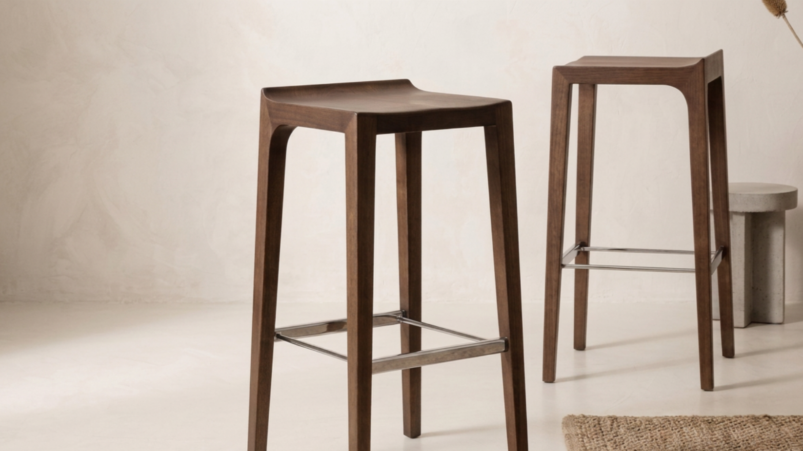 Two wooden bar stools, one with a slightly curved seat, are placed inside a room with a light-colored wall and minimal decor. In the background, there is a small round concrete side table.