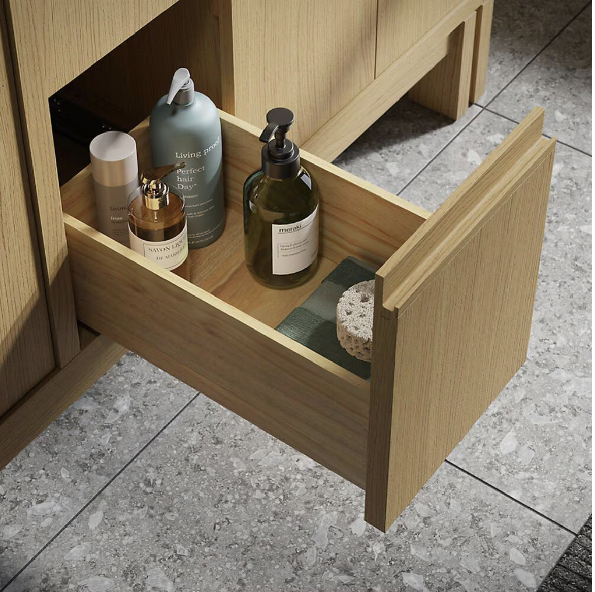 Open wooden bathroom vanity drawer containing skincare bottles, soap dispensers, towels, and a pumice stone