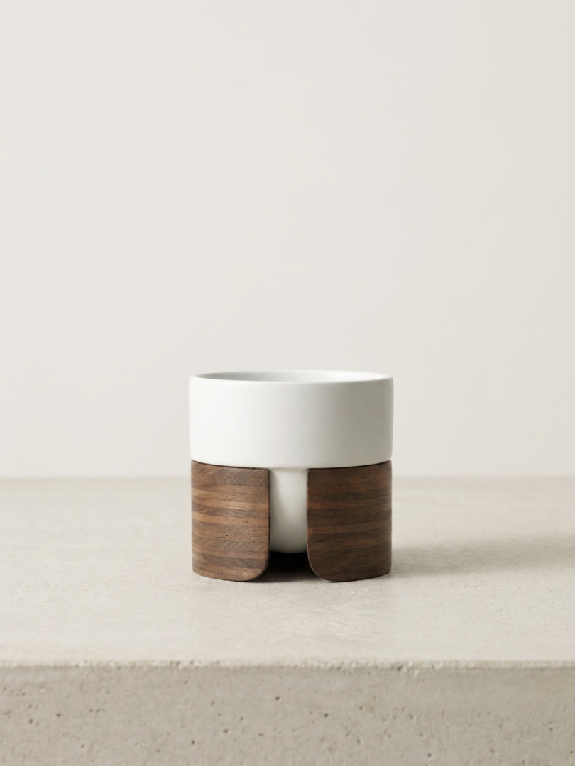 A white ceramic planter with a wooden stand on a light-colored surface against a plain off-white background.
