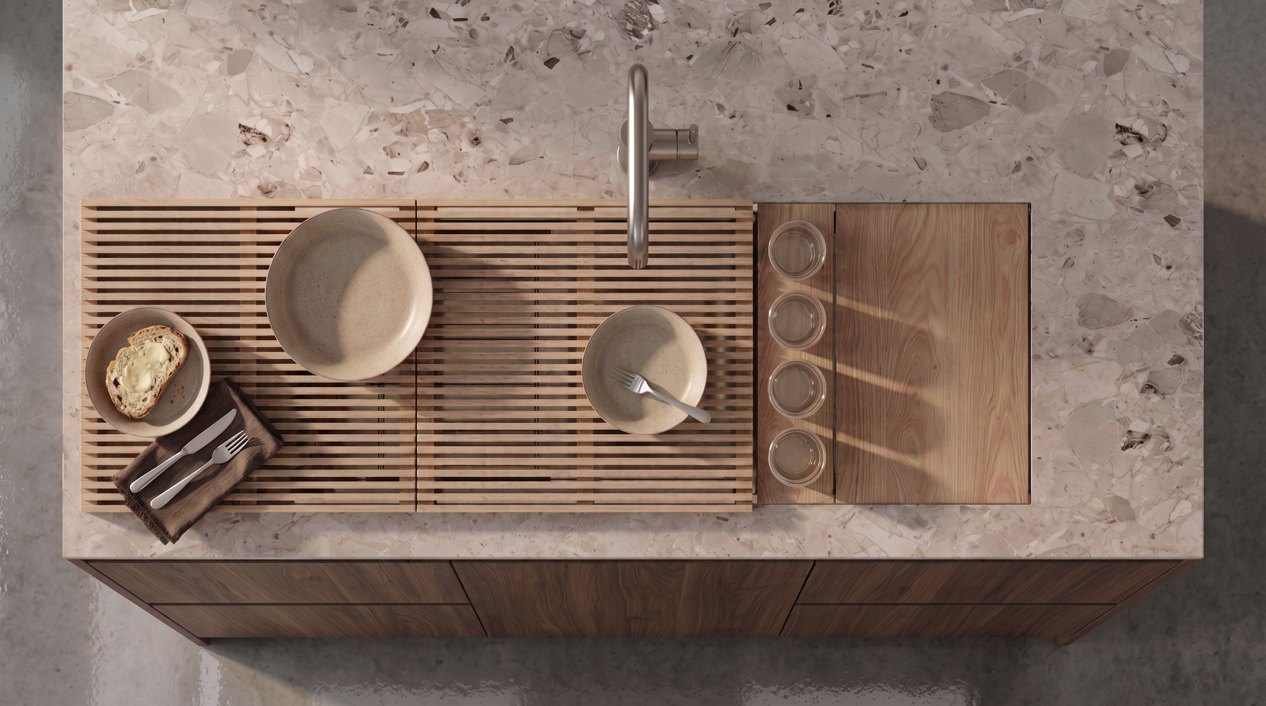 Top-down view of a kitchen counter with a wooden and bamboo placemat, a small bowl with a piece of bread and butter, an empty larger bowl, a smaller bowl with a fork, a folded napkin with knives and forks, and four glasses aligned on the right side.