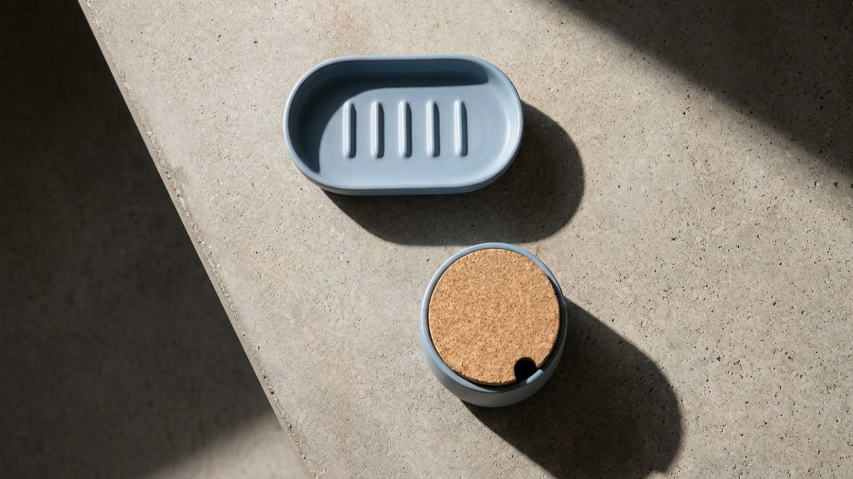 A light blue soap dish and a small round container with a cork lid on a beige countertop, casting shadows from sunlight.