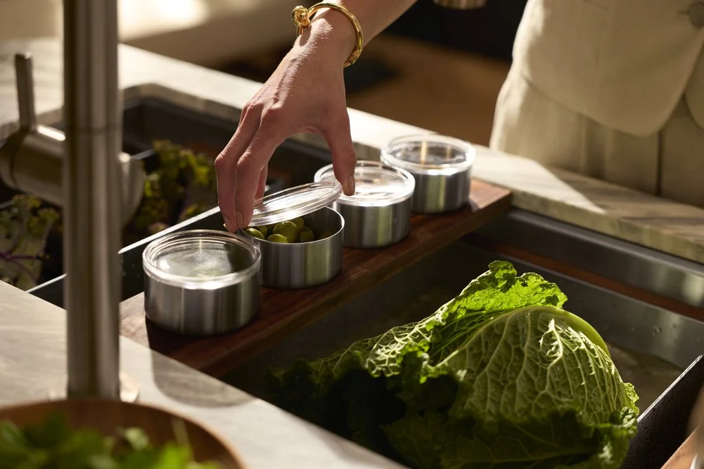 A person with a gold bracelet reaching to pick up a small tin container filled with green olives, among other similar containers, on a wooden tray. A large head of lettuce rests on a sink or counter nearby.