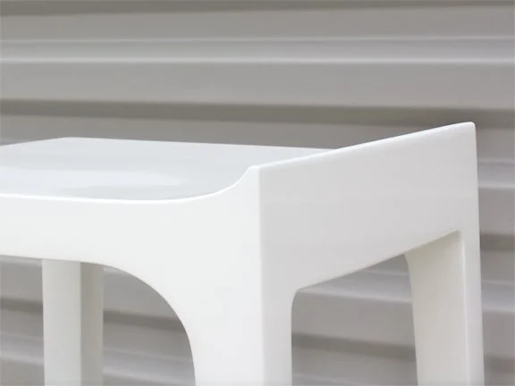 Close-up of a white plastic stool with a curved seat and straight legs against a gray background.