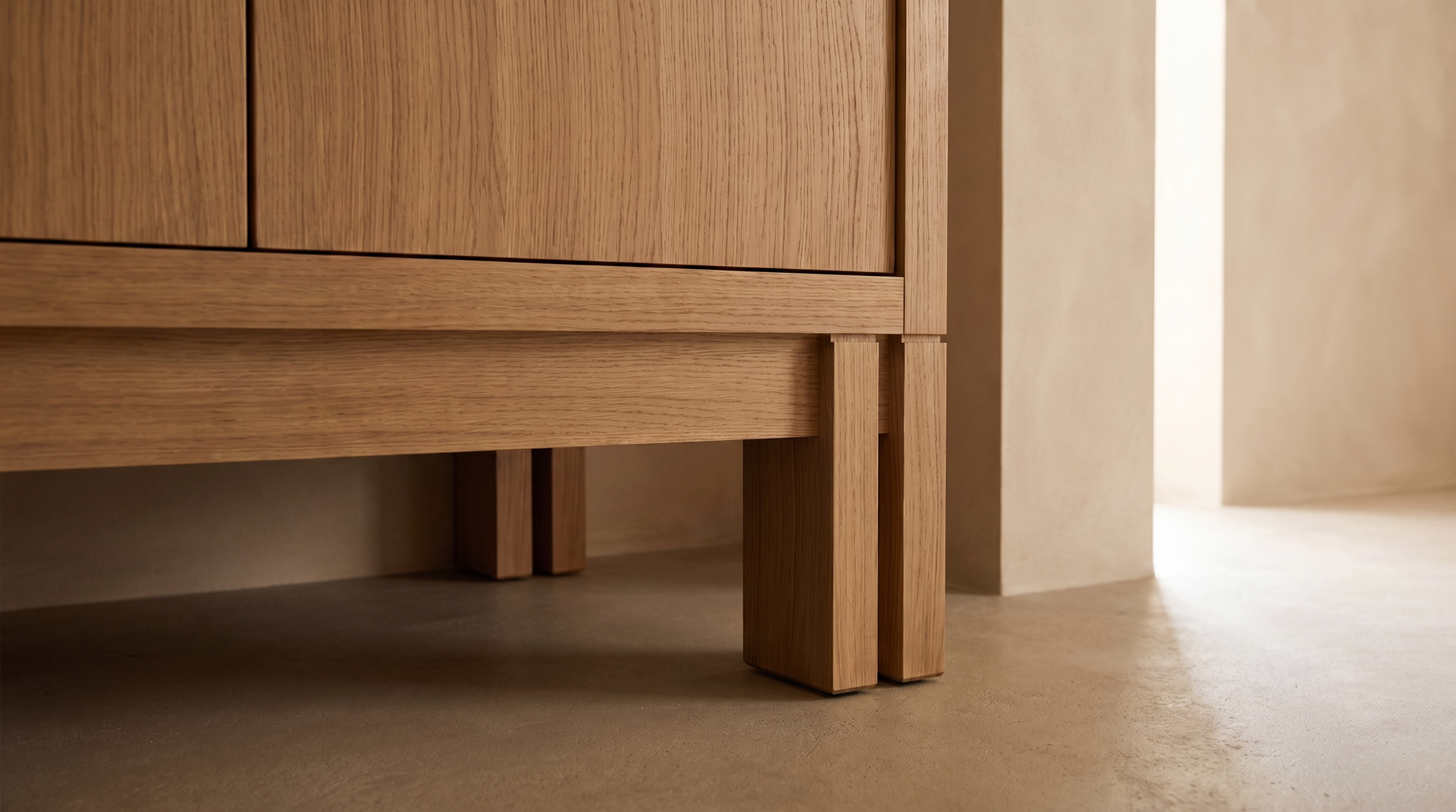 Close-up of a wooden cabinet or furniture piece with detailed wood grain, supported by thick wooden legs, placed on a beige carpeted floor next to a white wall.