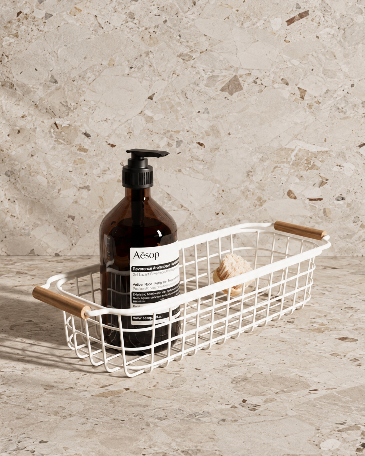 Aesop Reverence Aromatique Hand Wash in an amber pump bottle inside a white wire basket with wooden handles and a natural hand brush