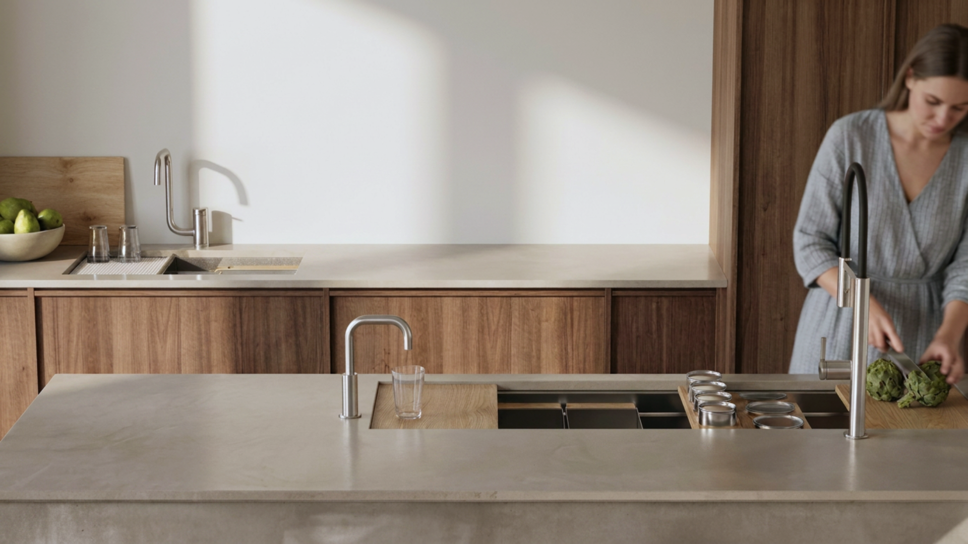 A woman in a gray dress is washing green vegetables at a kitchen sink with a black faucet, in a modern kitchen with wooden cabinets and light-colored countertops.
