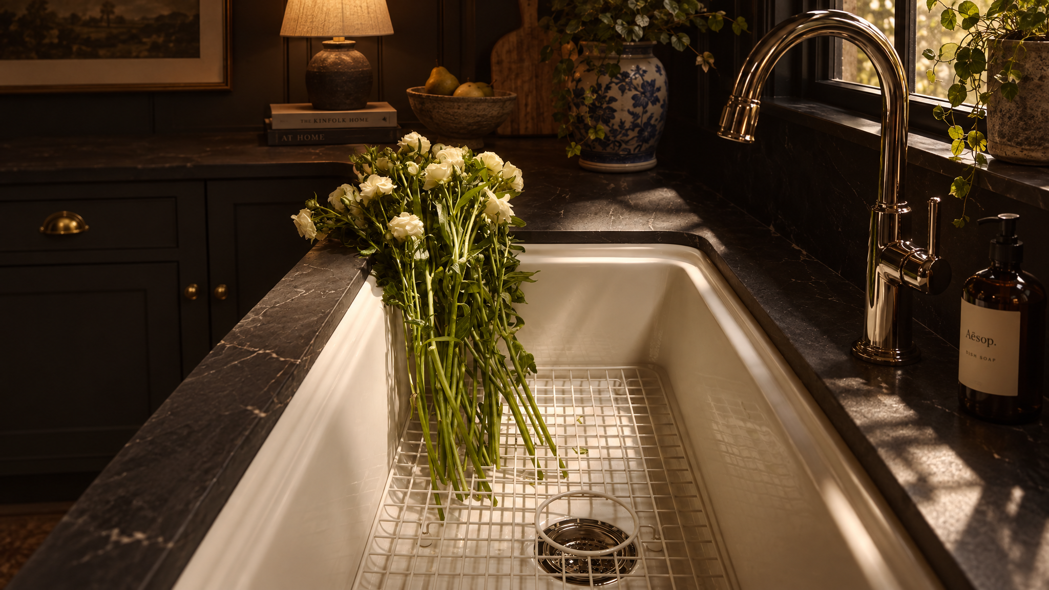 Elegant dark kitchen with white farmhouse sink, white flowers, brass faucet, marble countertops, and Aesop dish soap