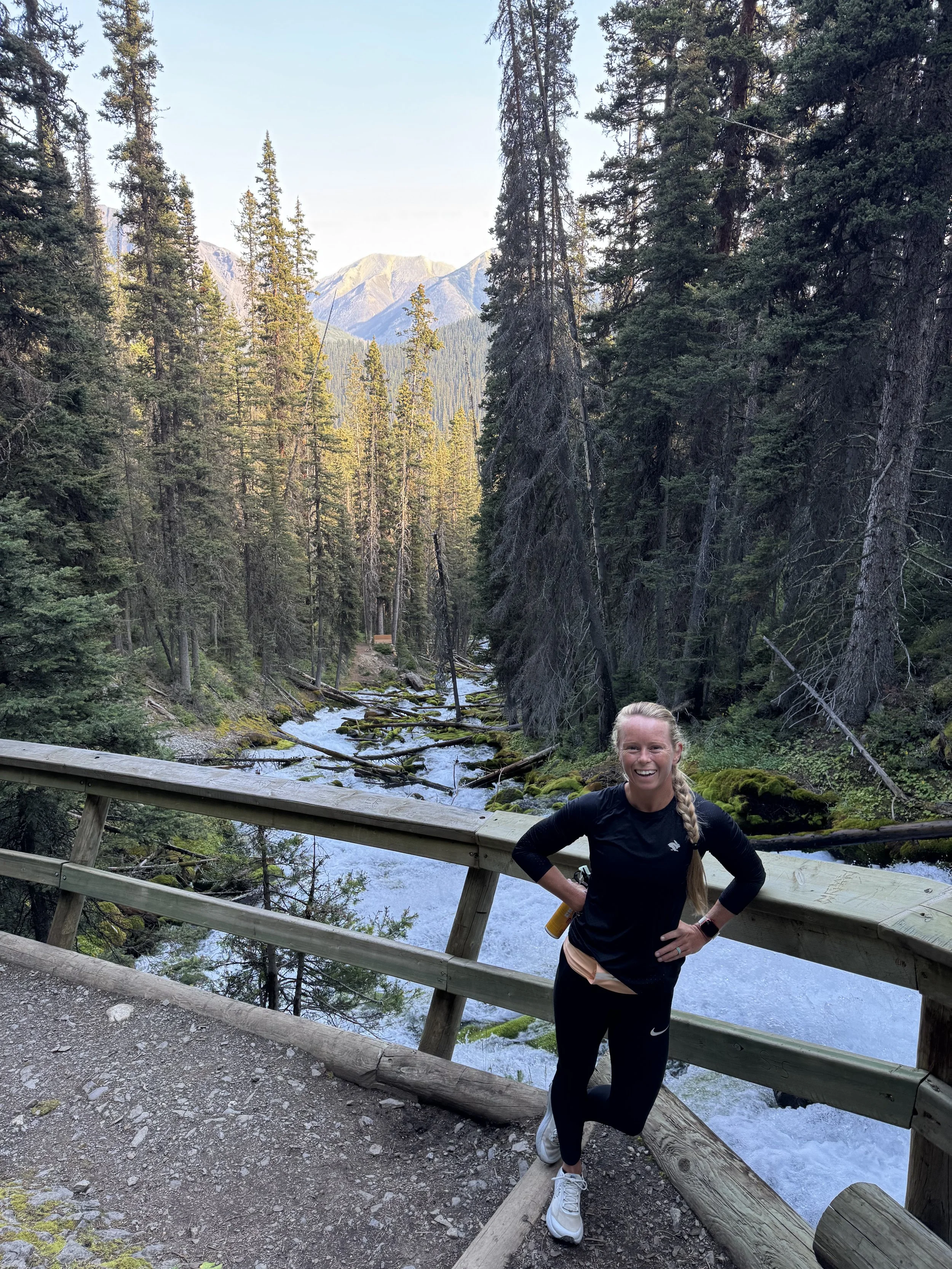 A woman with long blonde hair in a braid, wearing black athletic clothing and white sneakers, leans against a wooden railing on a trail in a forested mountainous area. She is smiling and has one hand on her hip. Behind her is a flowing river and tall evergreen trees, with mountains in the distance.