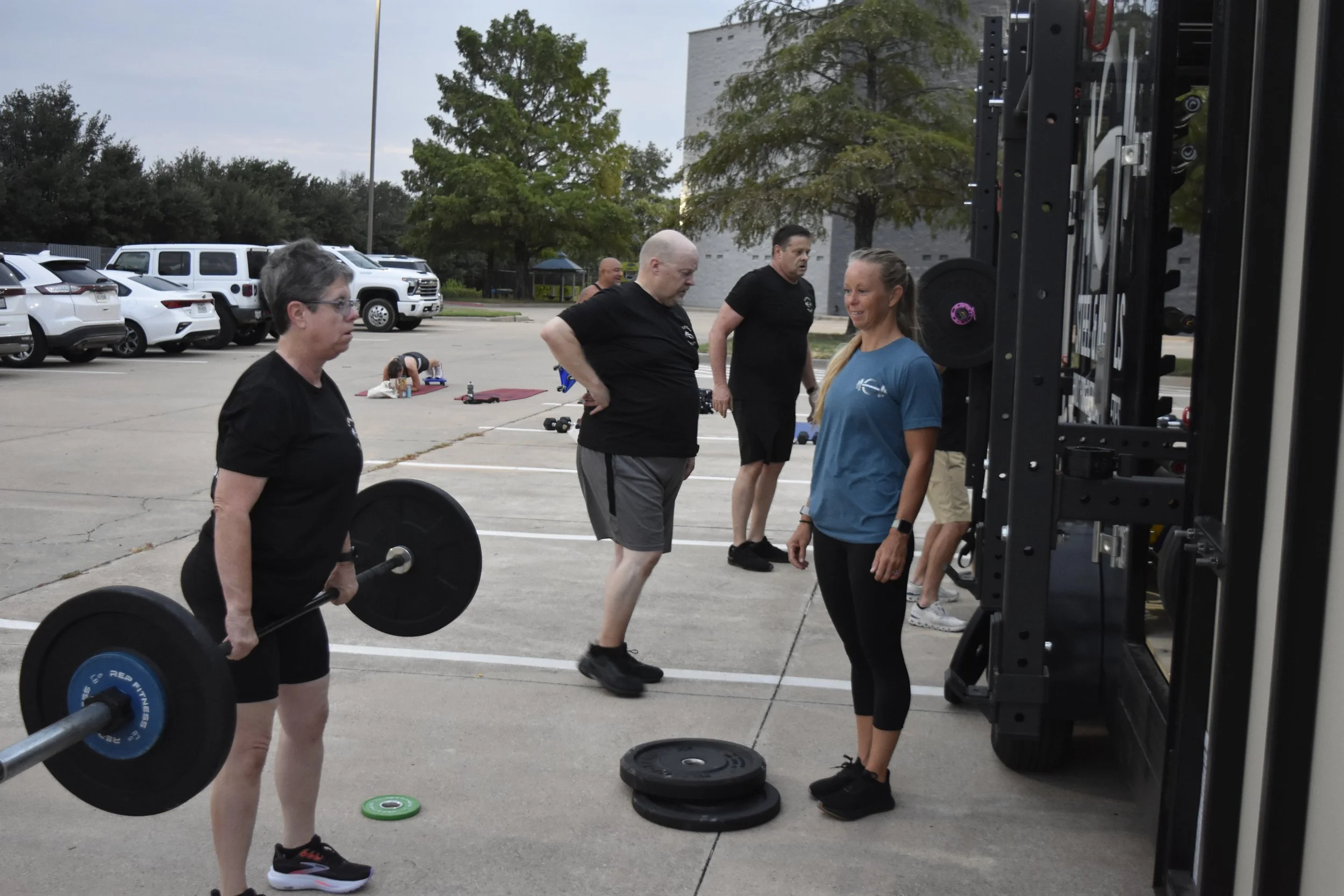 A woman lifting a barbell at an outdoor gym with a trainer.