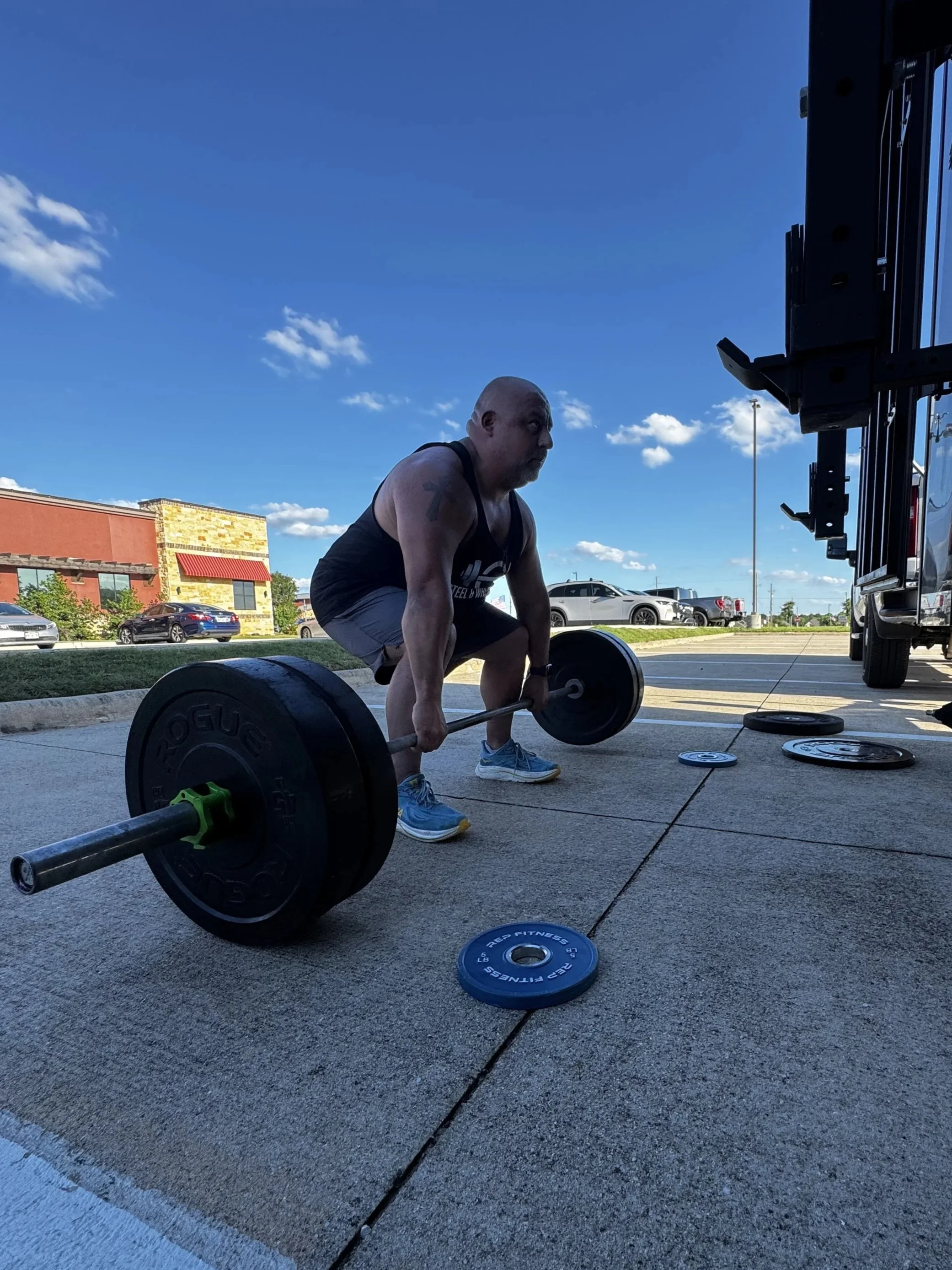 A bald man in a black tank top and gray shorts is bending over to lift a barbell with black weights outdoors in a parking lot. There are cars parked in the background and a building with a red awning. The sky is blue with some scattered clouds.