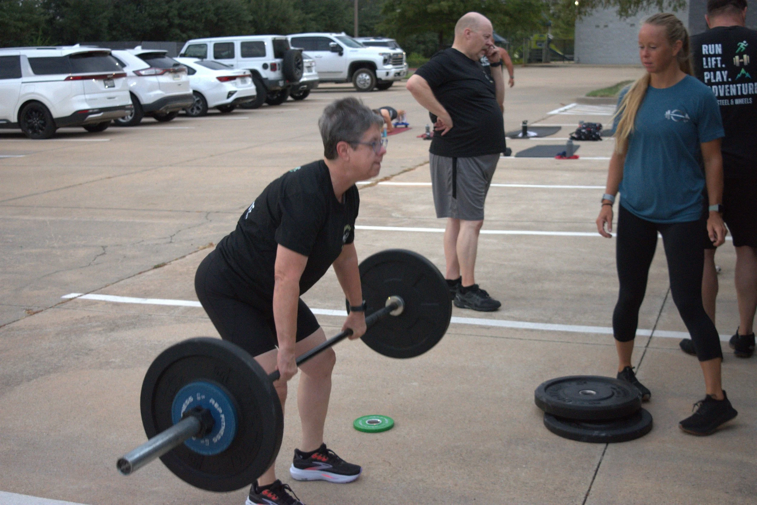 A woman lifting a barbell at an outdoor parking lot gym session while three other people, including a man with a shaved head and a woman in a blue shirt, observe. Several parked cars are visible in the background.